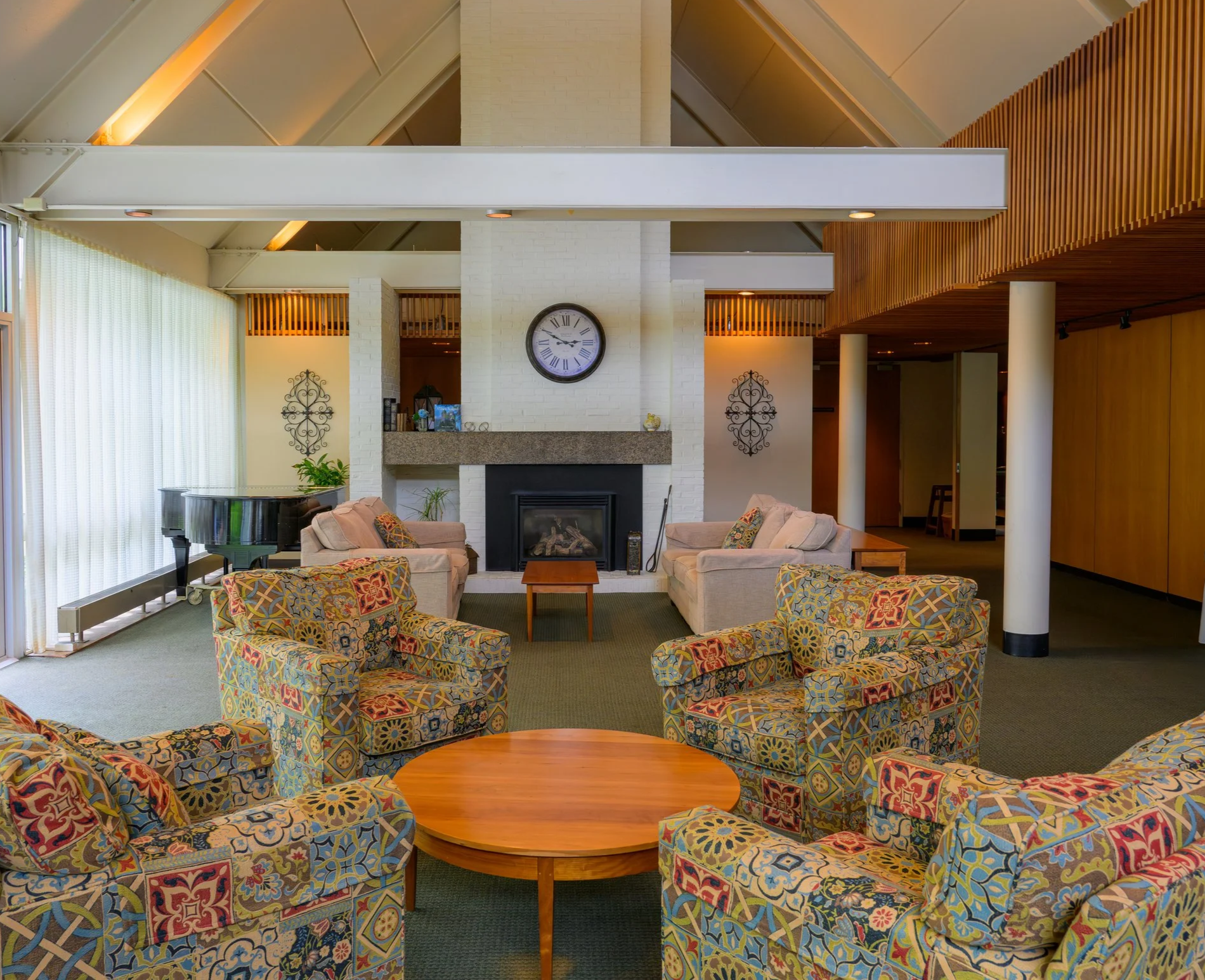 Interior shot of a cozy living room featuring a white brick fireplace with a clock above it, surrounded by cushioned armchairs with colorful patterns, a dark-colored grand piano, and decorative wall arts, with large windows covered by sheer curtains and a wooden ceiling design.