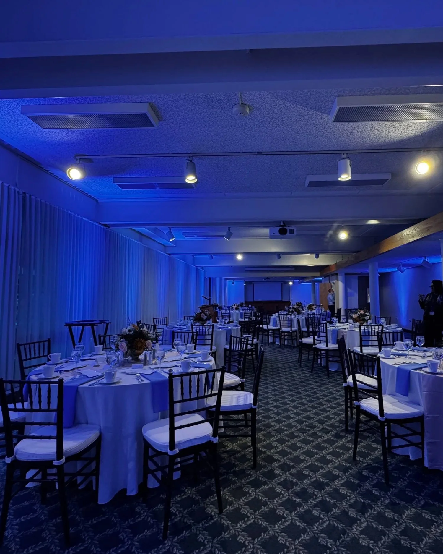 Wedding reception decor with round tables covered in white tablecloths, decorated with floral centerpieces, set with plates, glasses, and utensils, and surrounded by black chairs with white cushions, in a room illuminated with blue lighting and a pat