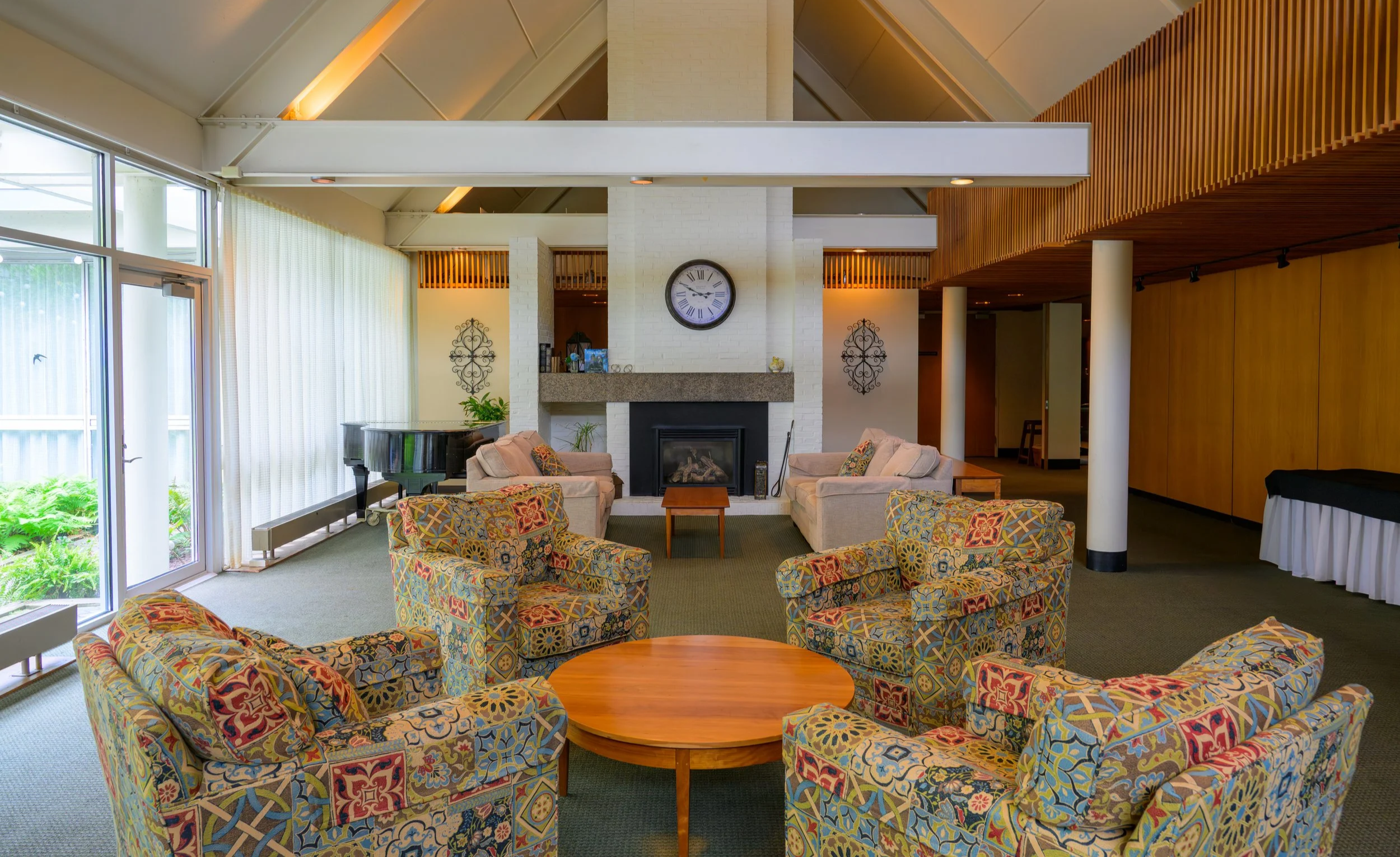 Interior of a living room with a white brick fireplace, a large round wall clock above it, a black grand piano to the left, colorful patterned armchairs and sofas around a wooden coffee table, large windows with white curtains on the left, wooden paneling on the ceiling and right wall, and decorative wall ornaments.