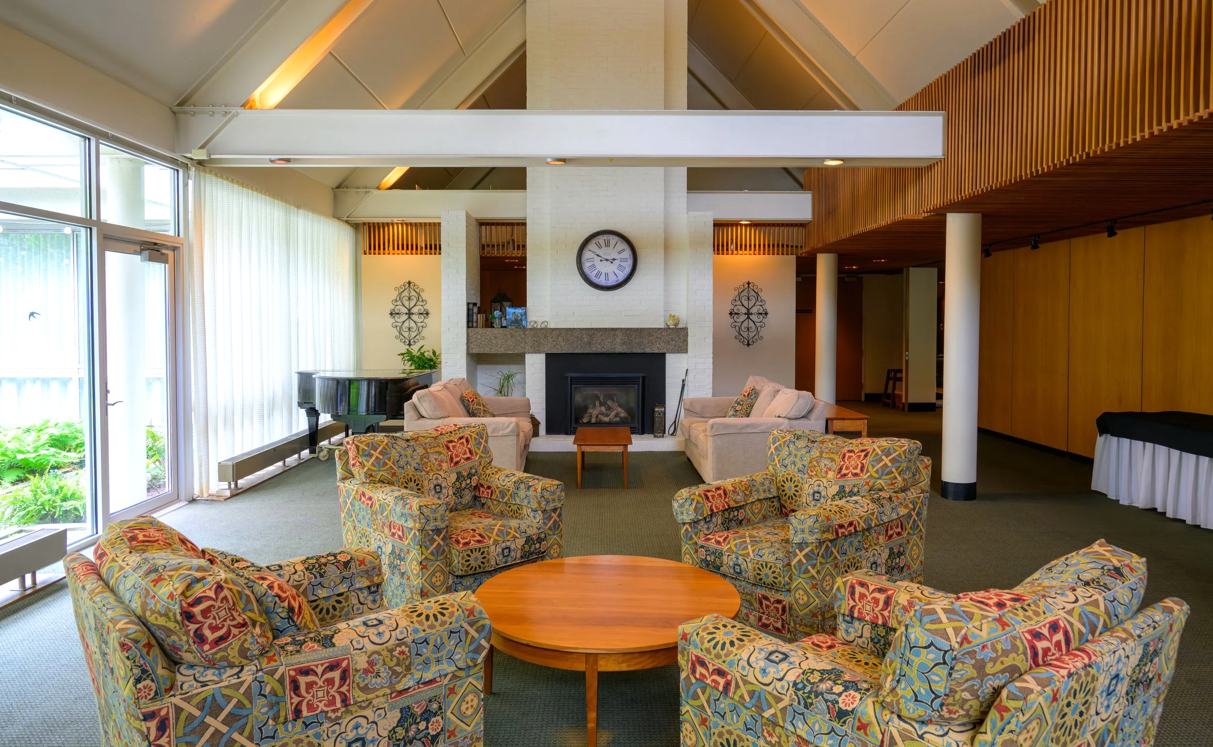 Interior of a living room with patterned armchairs arranged around a round wooden table, a white brick fireplace with a clock above it, large windows with curtains, and wooden accents on the ceiling and walls.