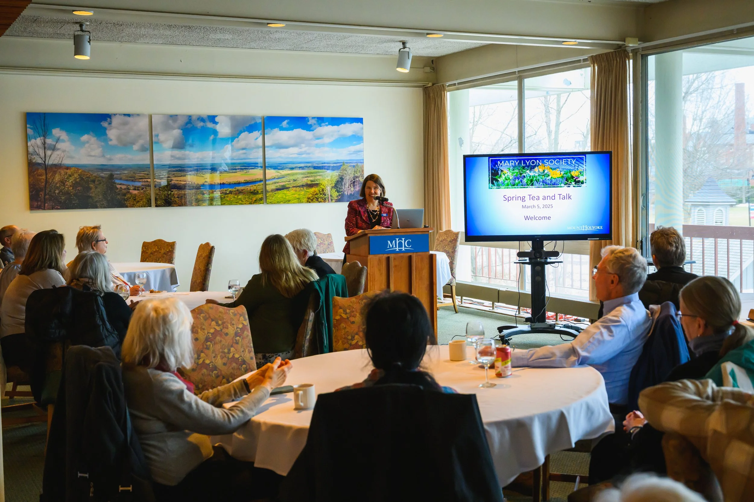 A woman giving a presentation at a peaceful indoor event with a large digital screen that reads 'Mary Lyon Society Spring Tea and Talk, March 5, 2025, Welcome,' and a scenic landscape painting on the wall behind her, audience members seated around tables, some with cups, in a room with large windows and beige curtains.