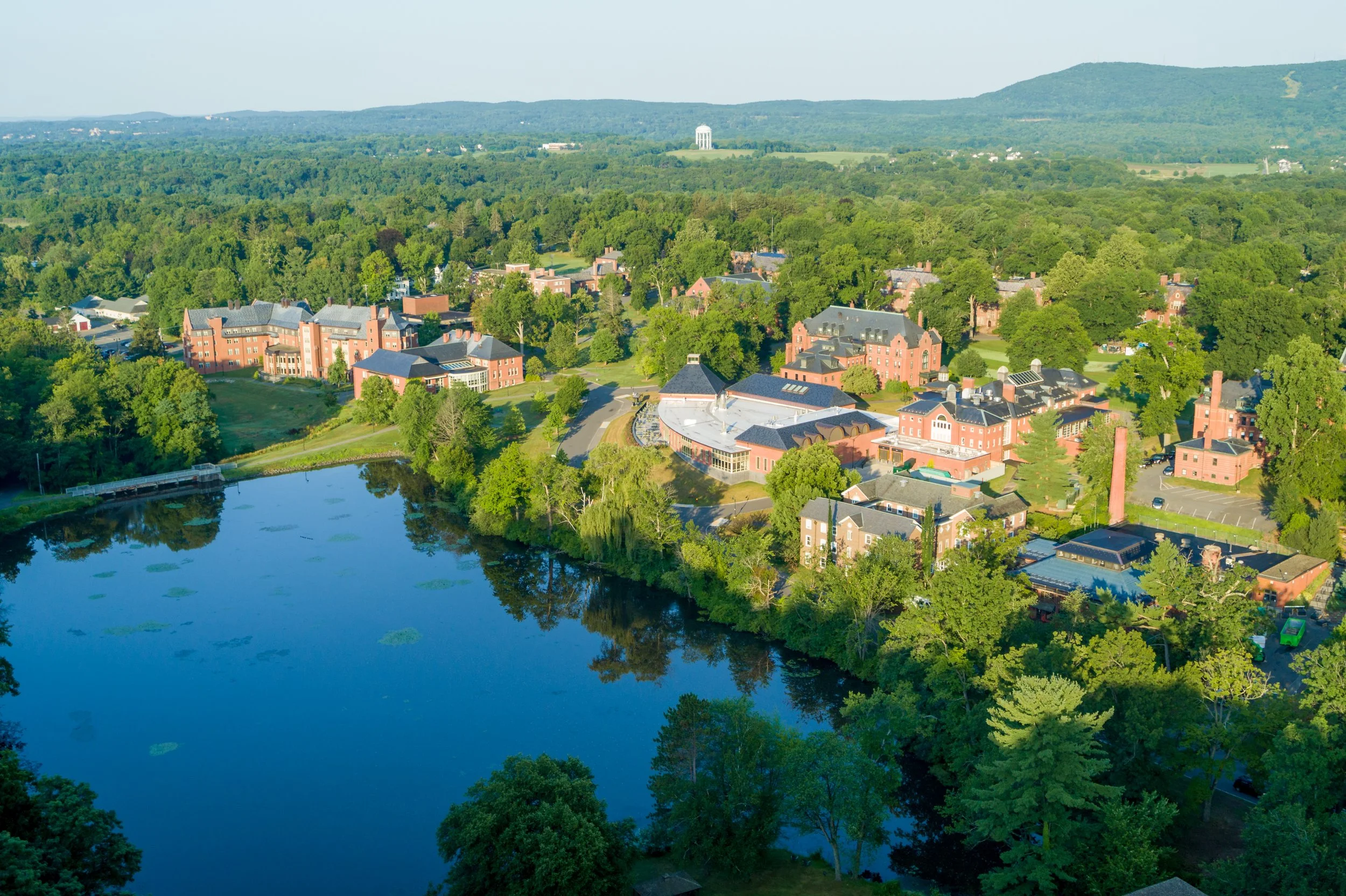 Aerial view of a college campus surrounded by lush green trees, with a pond in the foreground and mountains in the background.