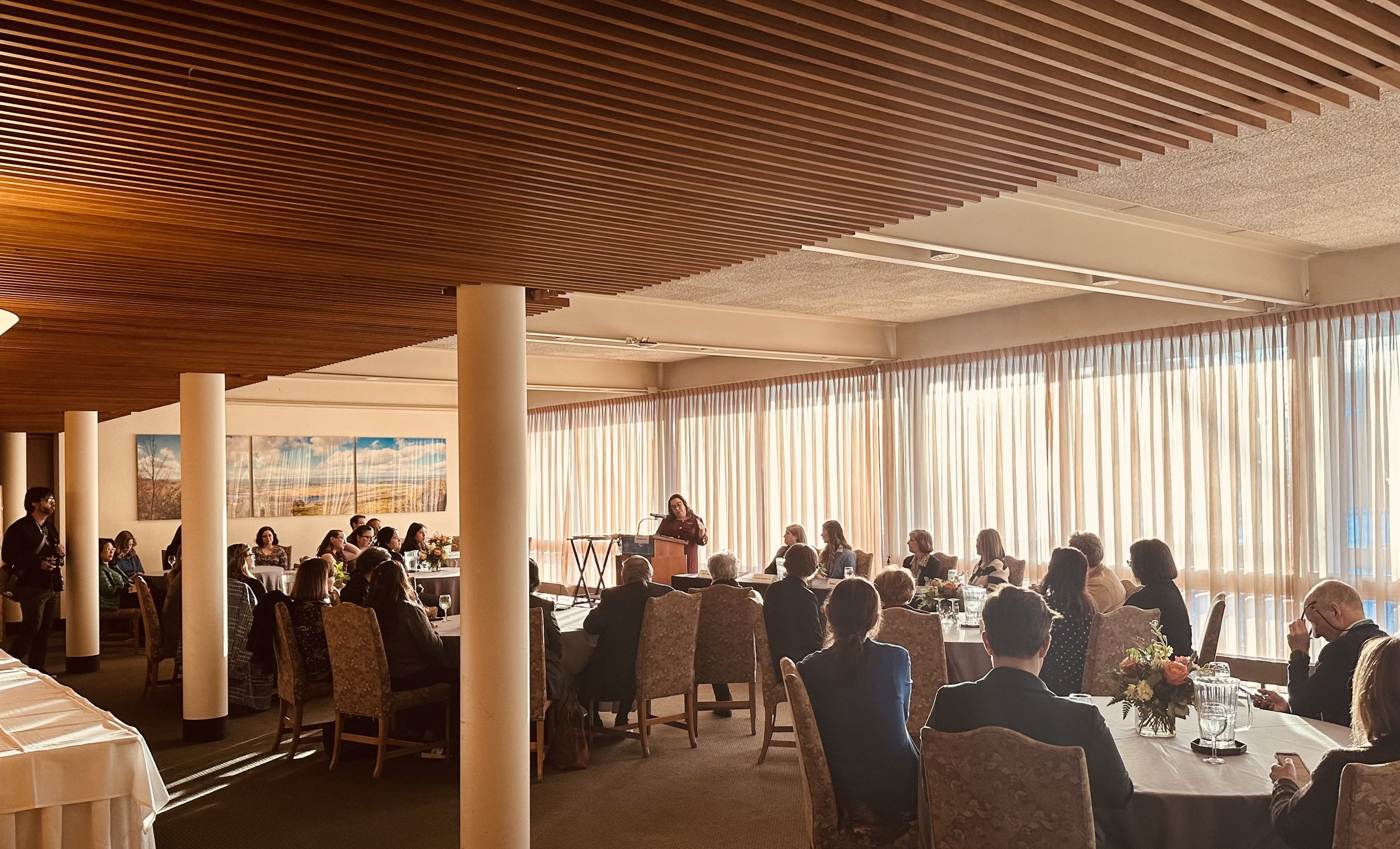 People attending a formal event or conference in a bright, spacious room with large windows and curtains. There is a woman speaking at a podium, and attendees are seated at tables.