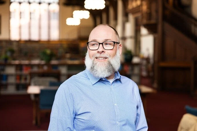A smiling middle-aged man with glasses and a gray beard standing in a cozy, warmly lit library or study area with bookshelves and warm lighting in the background.