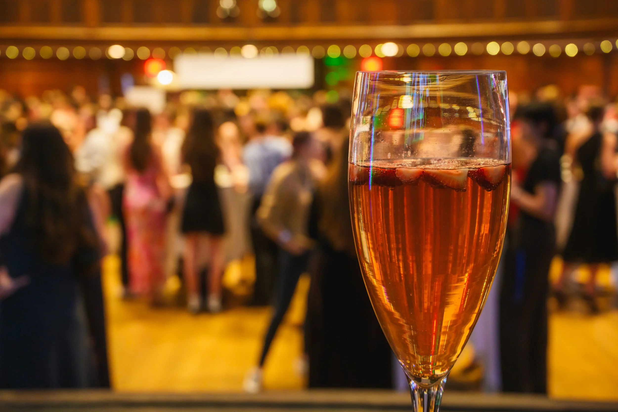 A glass of rosé wine with ice cubes against a background of a crowded party or event with blurry people and warm lighting.