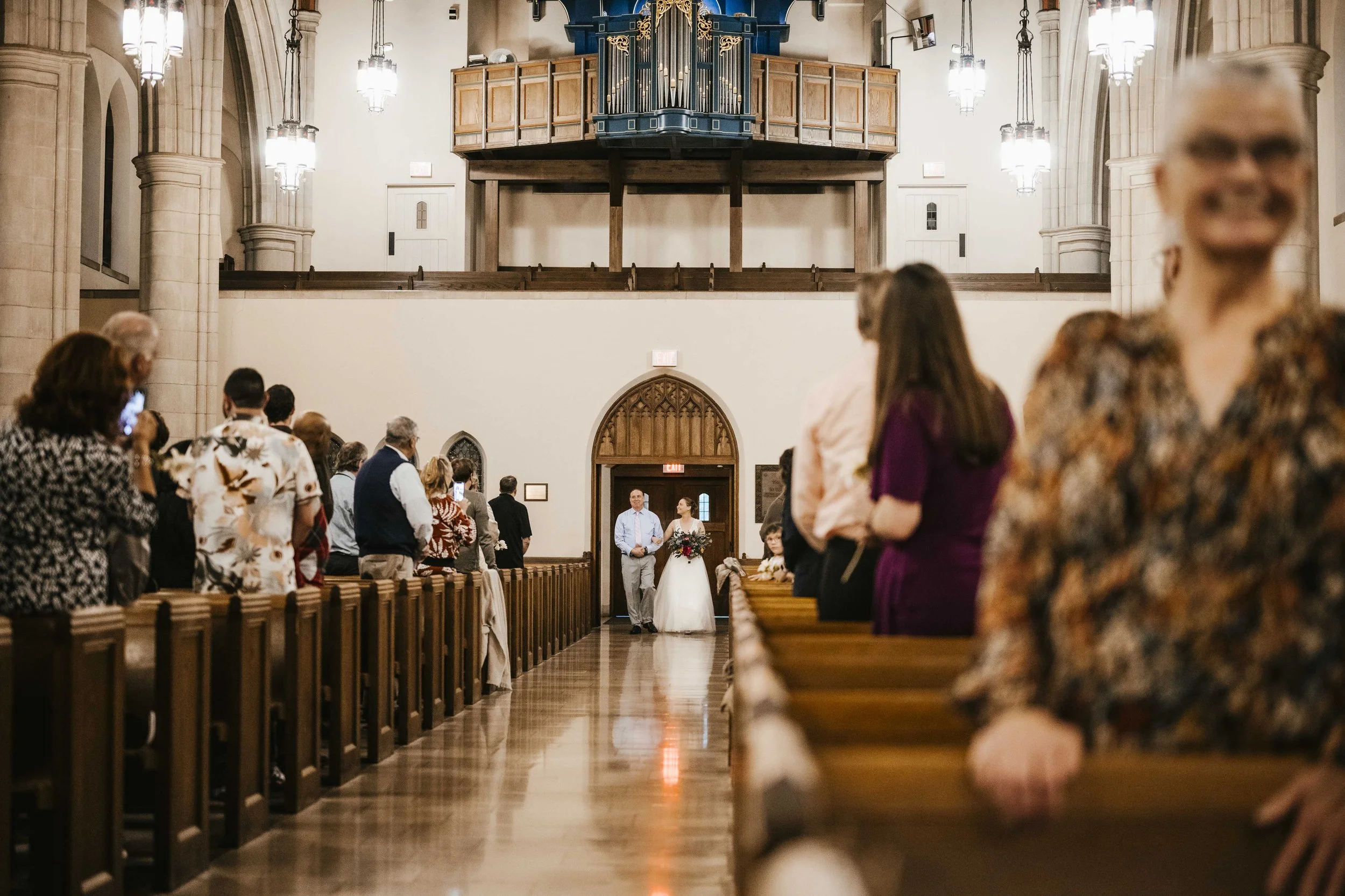 A wedding ceremony inside a church with guests seated on pews, a bride and groom at the altar, and a large pipe organ in the background.