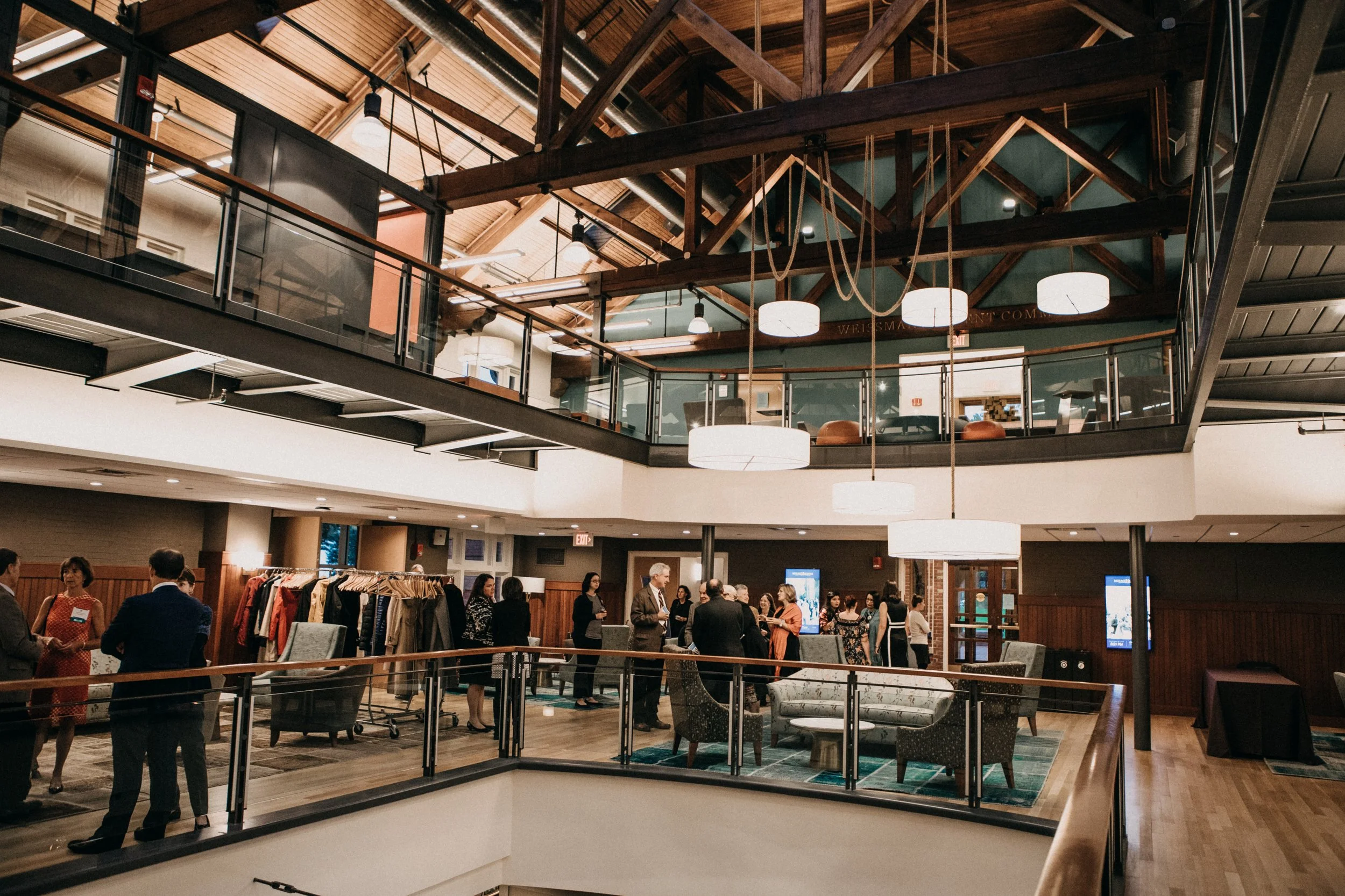 A group of people socializing and browsing clothing at a busy indoor event in a space with high wooden ceilings and modern lighting.