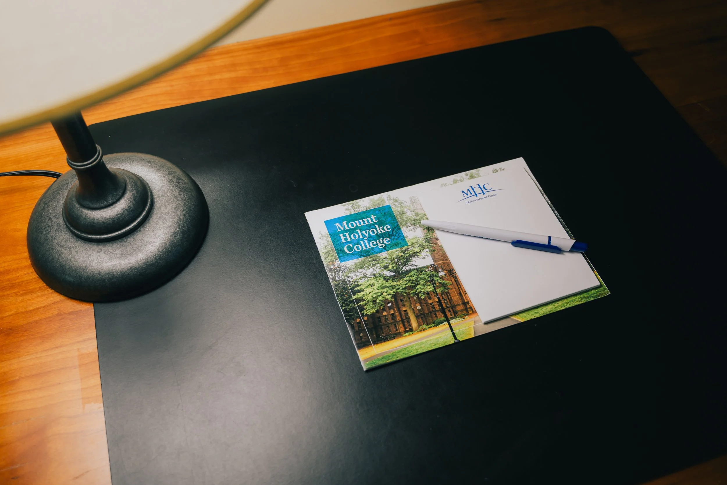 Desk with a lamp, a brochure from Mount Holyoke College, a notepad, and a pen.