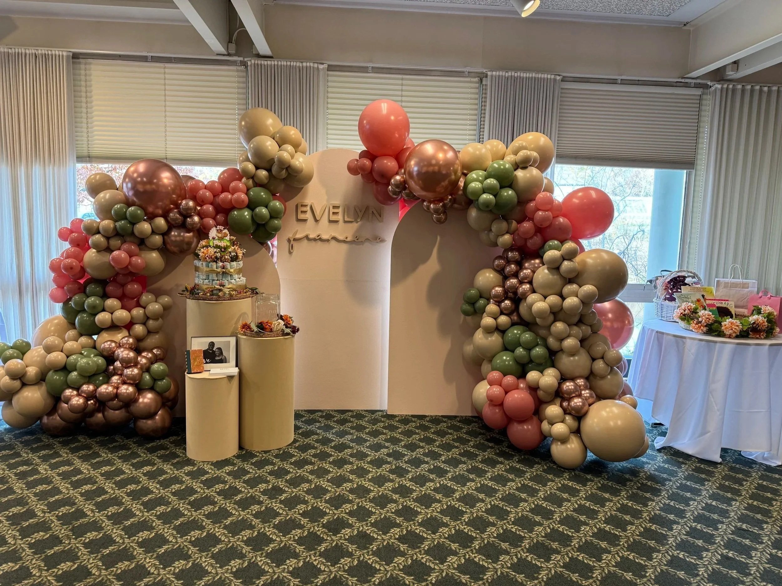 Decorative arch made of pink, cream, green, and metallic balloons surrounds a white backdrop with the name "EVELYN" and the word "party" on it. A small table with flowers and a basket are nearby, on a carpeted floor with a patterned black and yellow 