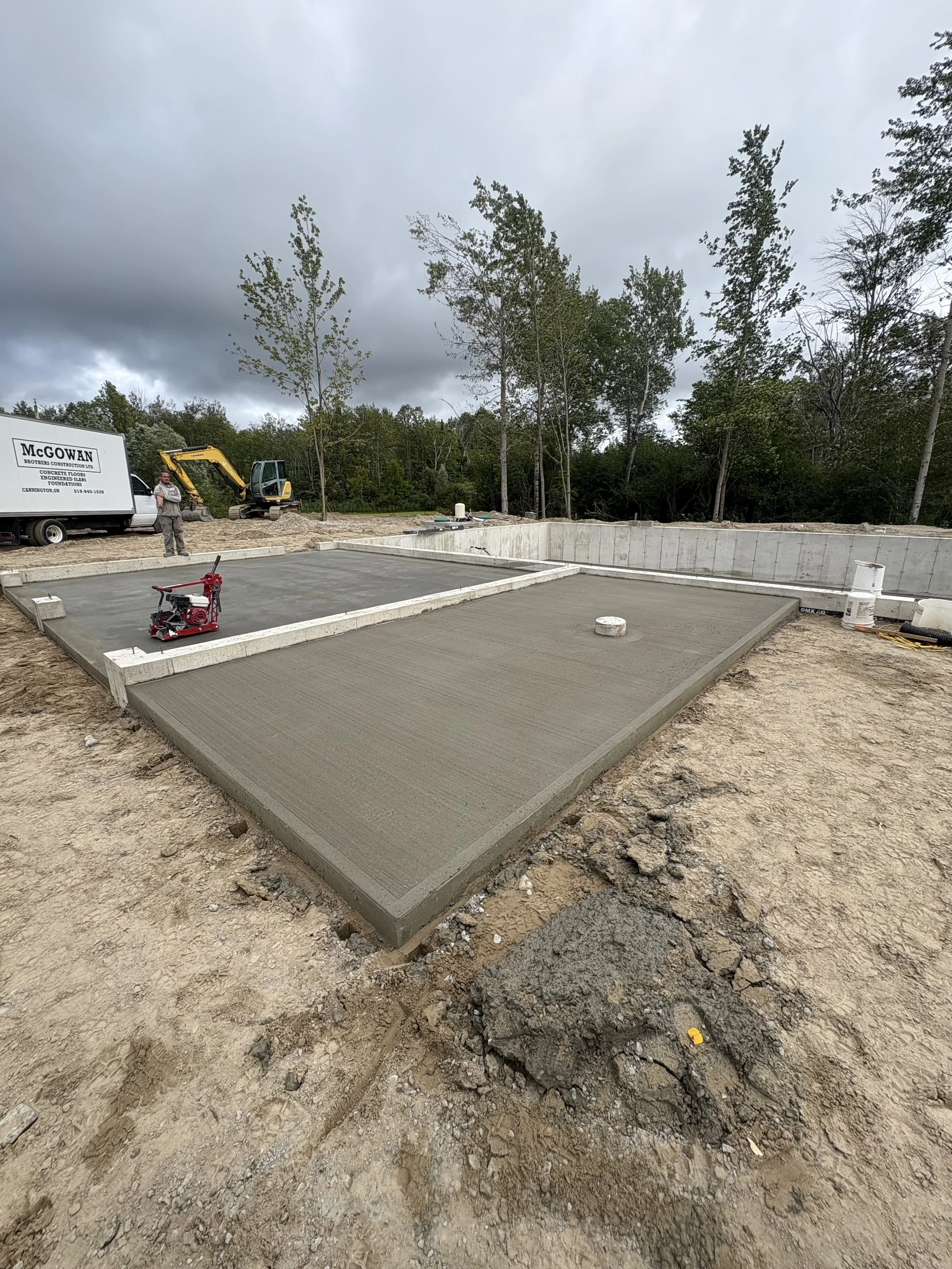 A partially constructed concrete foundation at a building site in Bracebridge with a small excavator, a person standing nearby, and trees in the background under a cloudy sky.