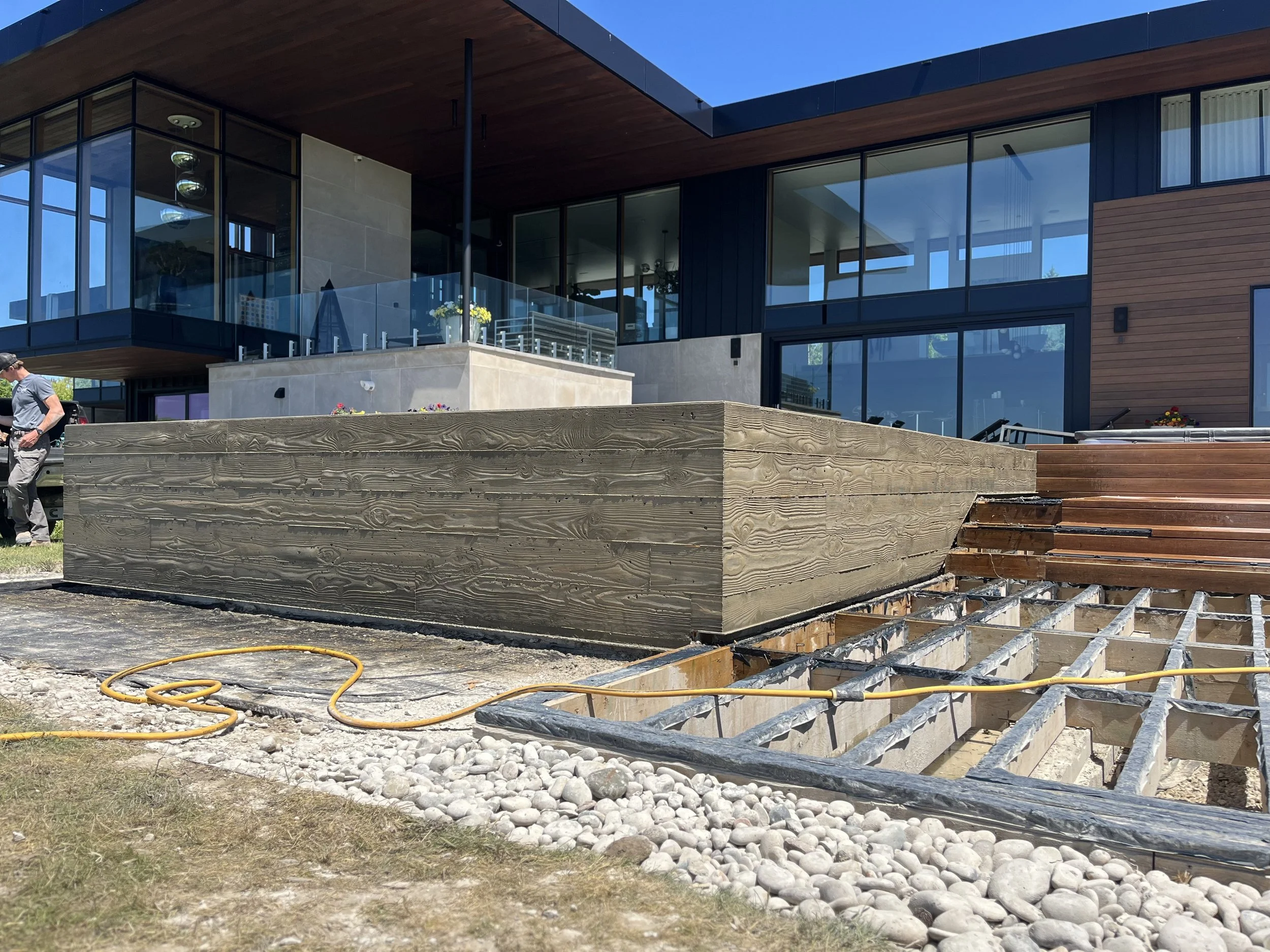 Modern house under construction with large glass windows, wooden accents, and a deck. Construction materials and tools are visible in the foreground, with a person walking on the site.
