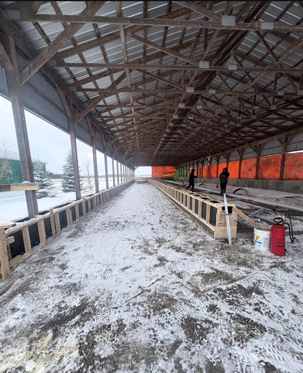 Construction site of an indoor sports facility with a snow-covered ground, showing a wooden structure and two workers installing or working on a raised platform.