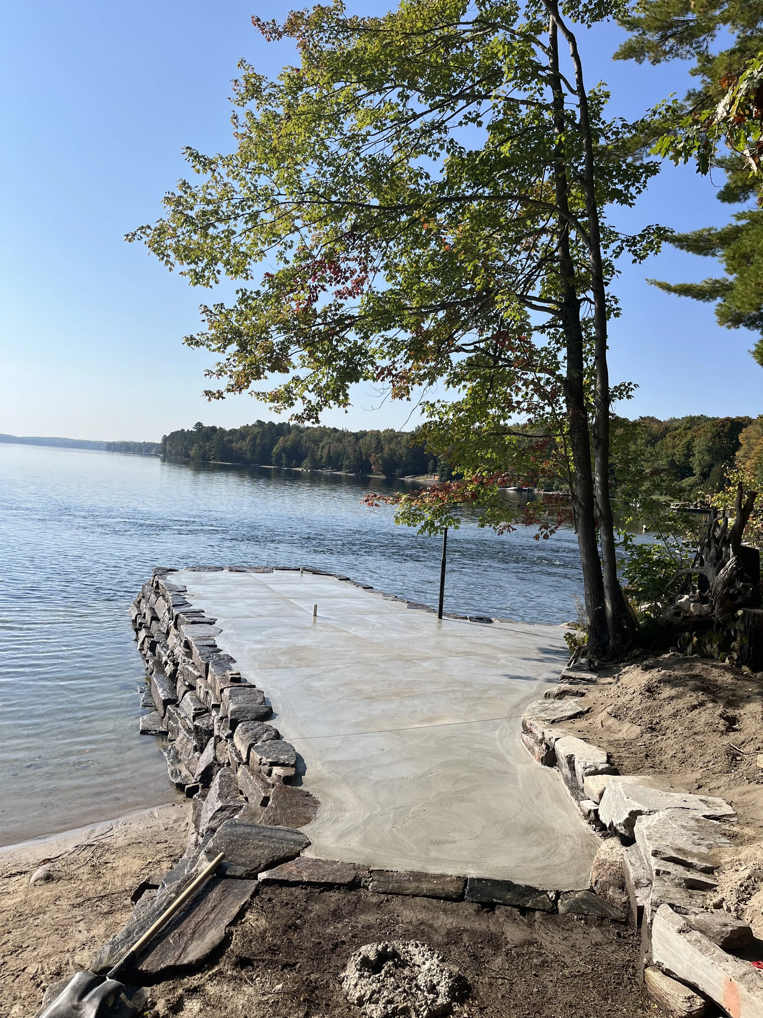A lakeside scene with a paved walkway extending into the water, bordered by stacked stones. There are trees with green leaves along the shoreline and a calm lake under a clear blue sky.