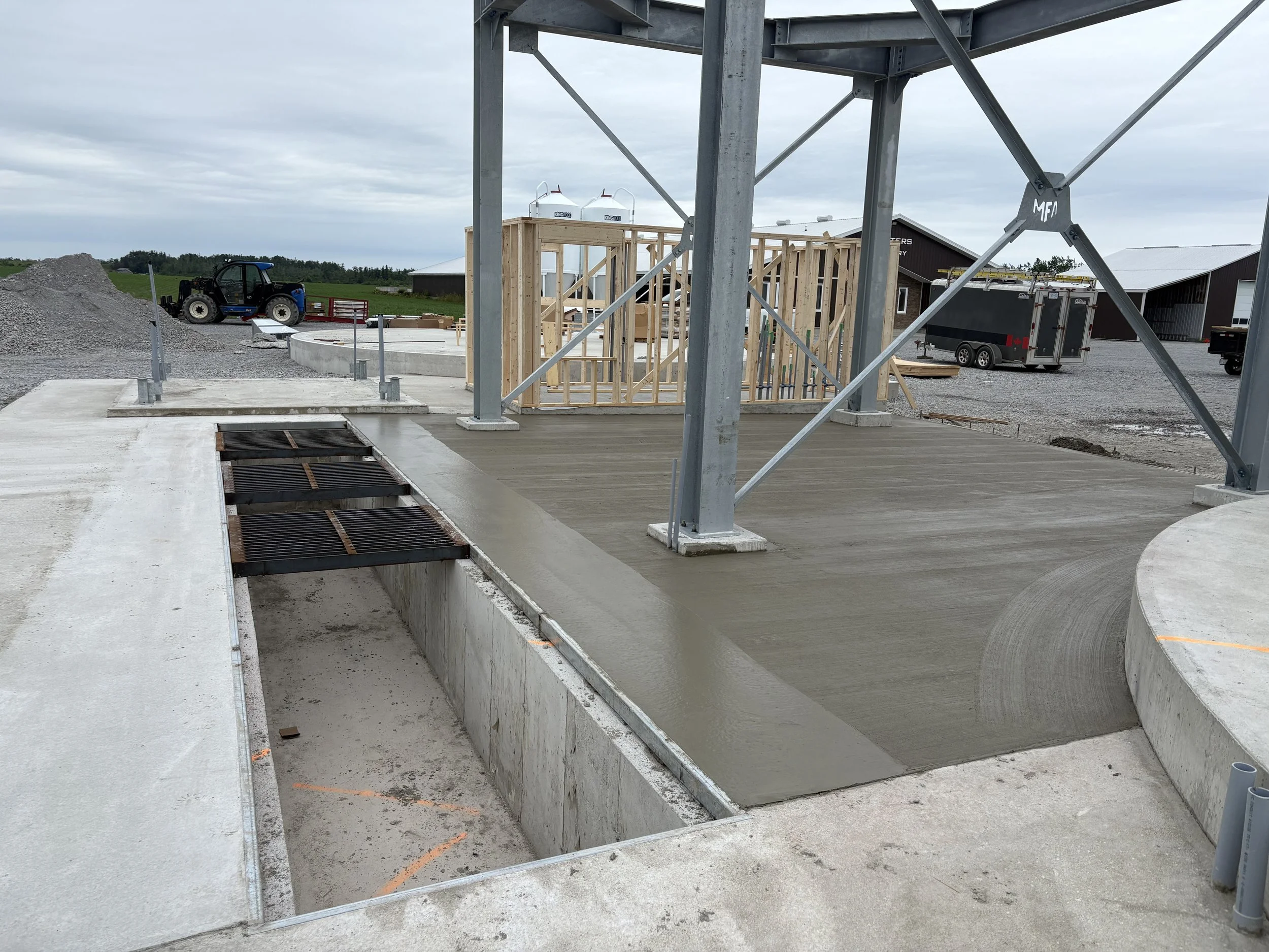 Construction site with partially built wooden framing, concrete foundation, and metal support structures, under a cloudy sky with nearby equipment and buildings.