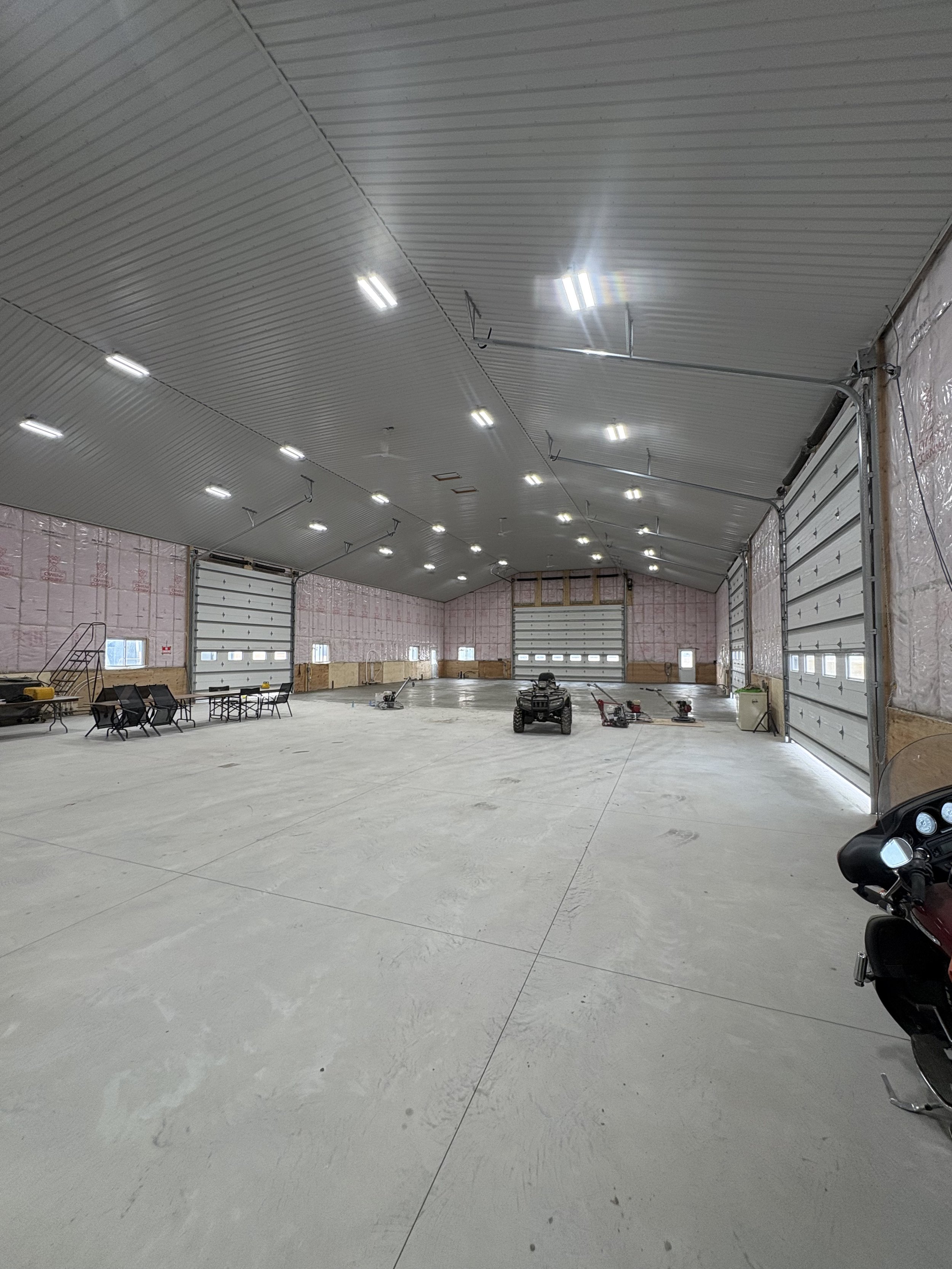 Interior of an unfinished large garage with a metal ceiling, concrete floor, multiple garage doors, and construction tools and equipment.