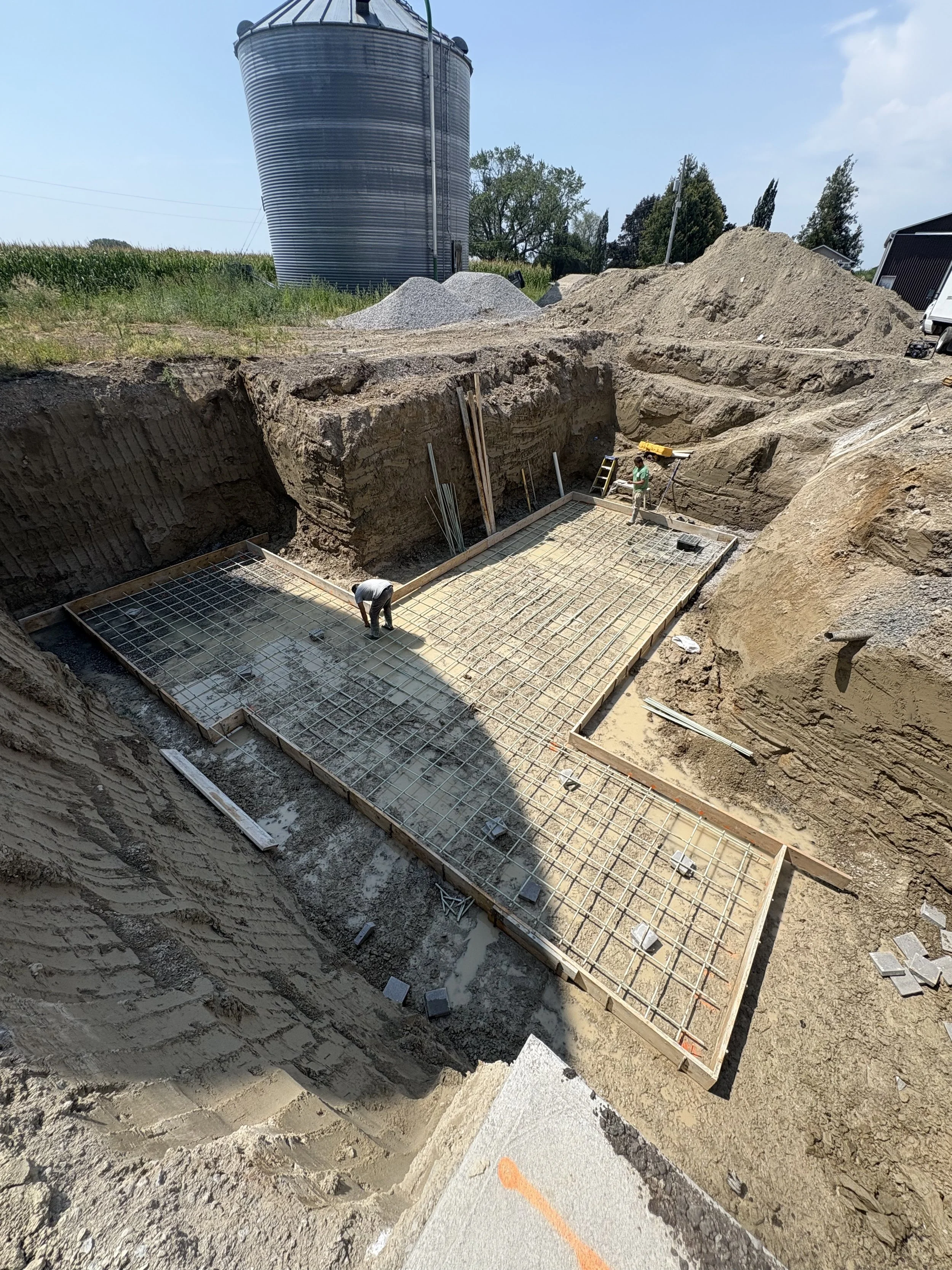 Construction site in Brock Township with a large excavation hole, concrete foundation framework, and two workers installing rebar.