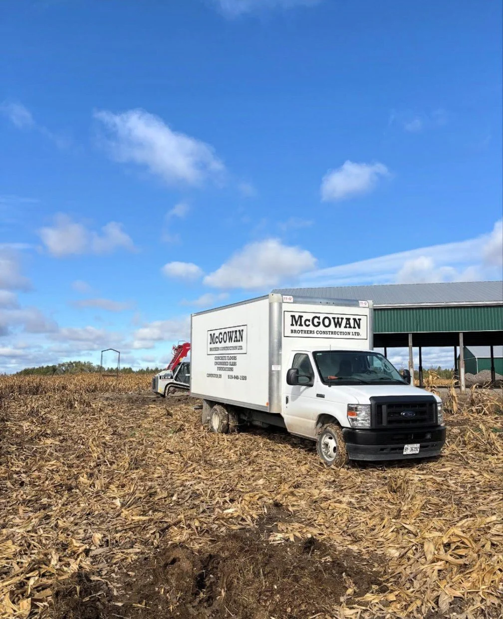 A white moving truck labeled McGowan Brothers Construction Ltd. parked in a harvested cornfield with a barn structure nearby, under a blue sky with scattered clouds.