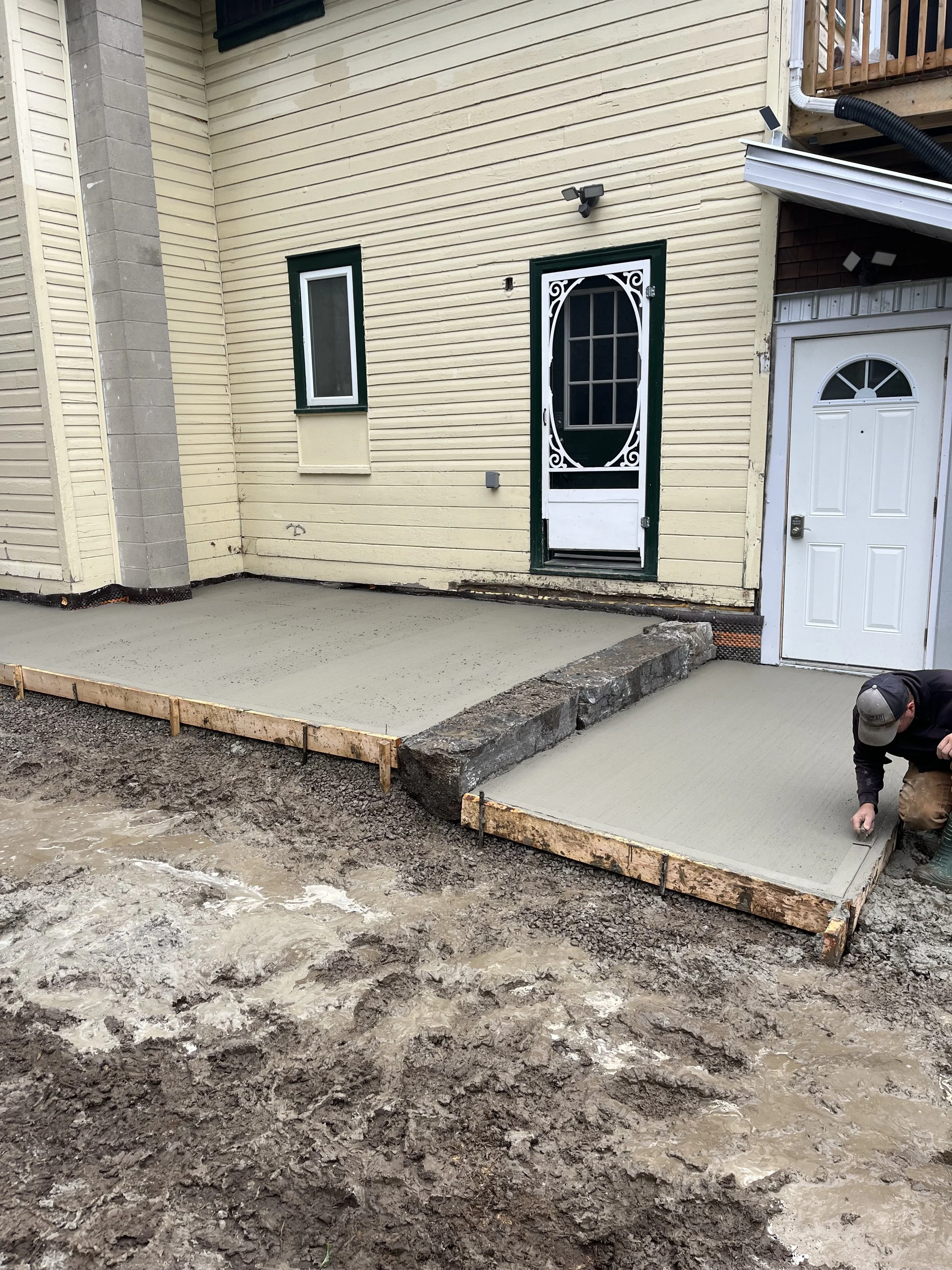 A construction worker is smoothing freshly poured concrete on a patio in a backyard in Oshawa, with a house featuring a window and a door in the background.