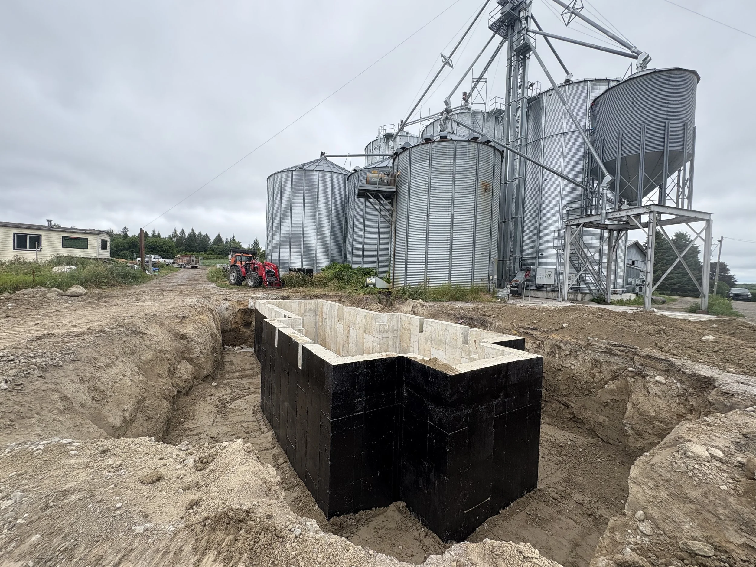 Construction site in Cannington with large metal grain silos, a red tractor, and an excavated foundation in the foreground under a cloudy sky.