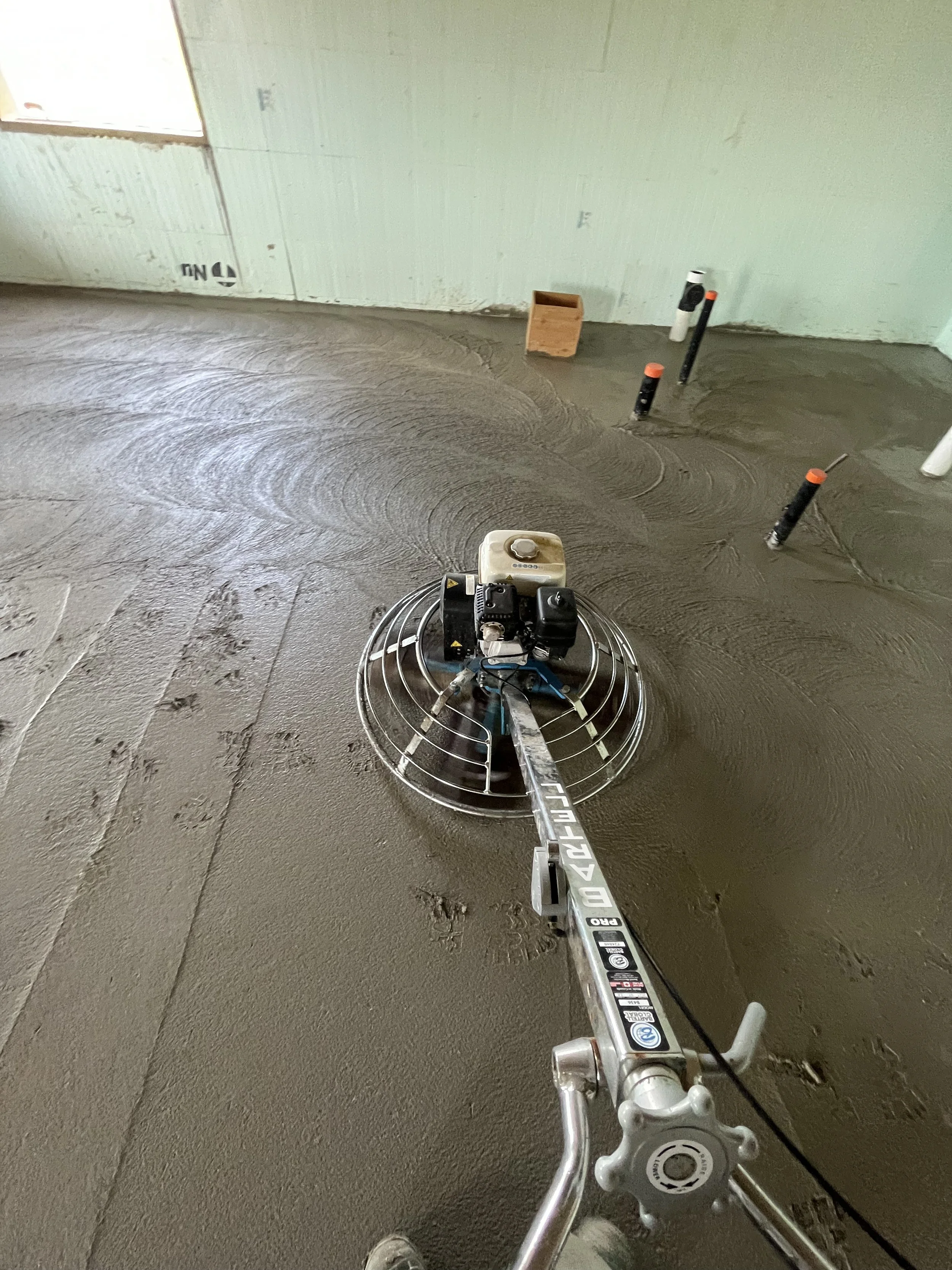 View of a concrete floor in a construction site with a floor grinding machine, a wooden block, and pipes protruding from the floor.