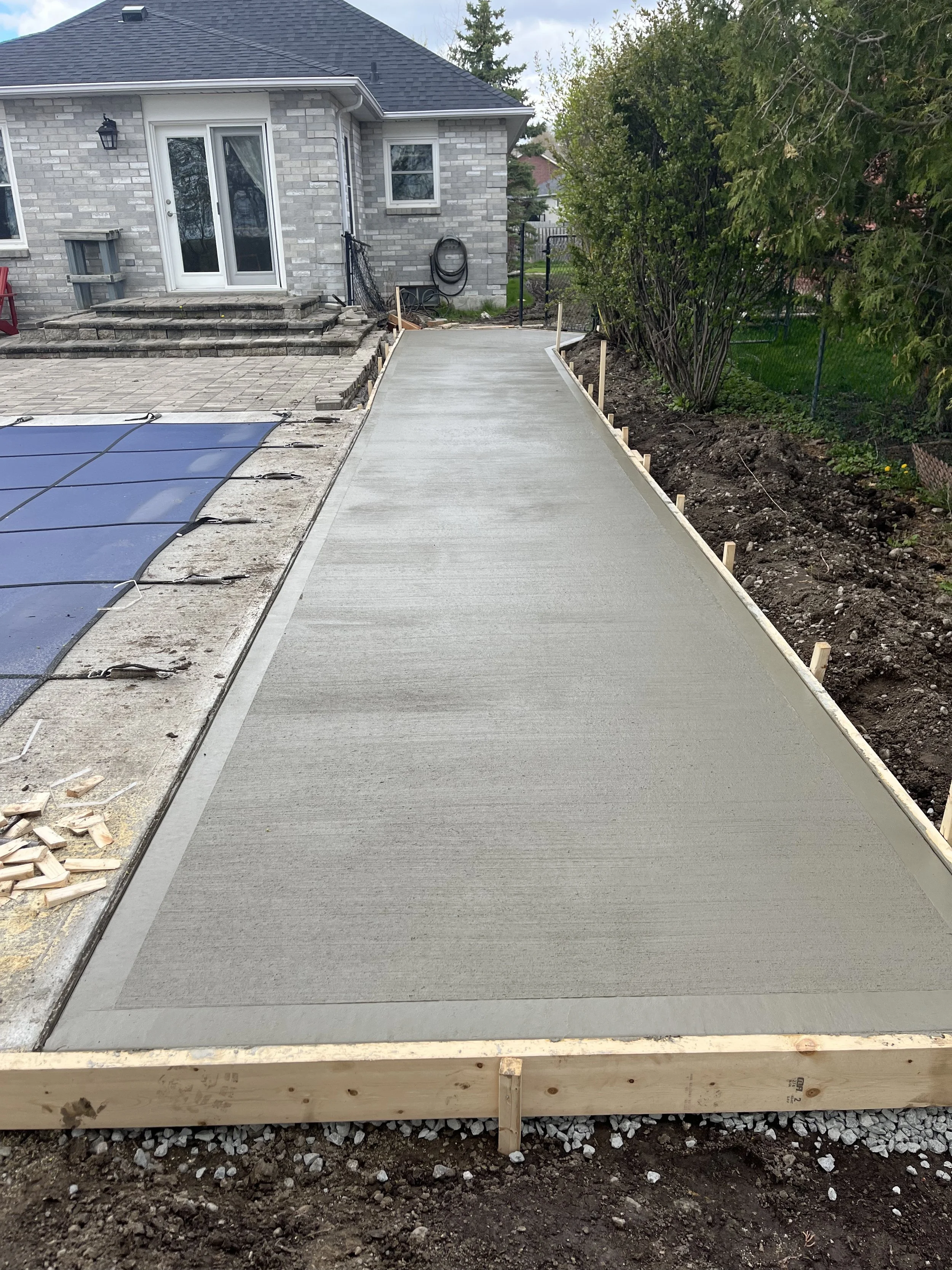 Newly poured concrete sidewalk with wooden framing, leading to a house with a brick exterior, sliding glass door, and outdoor staircase, surrounded by a garden with trees and soil.