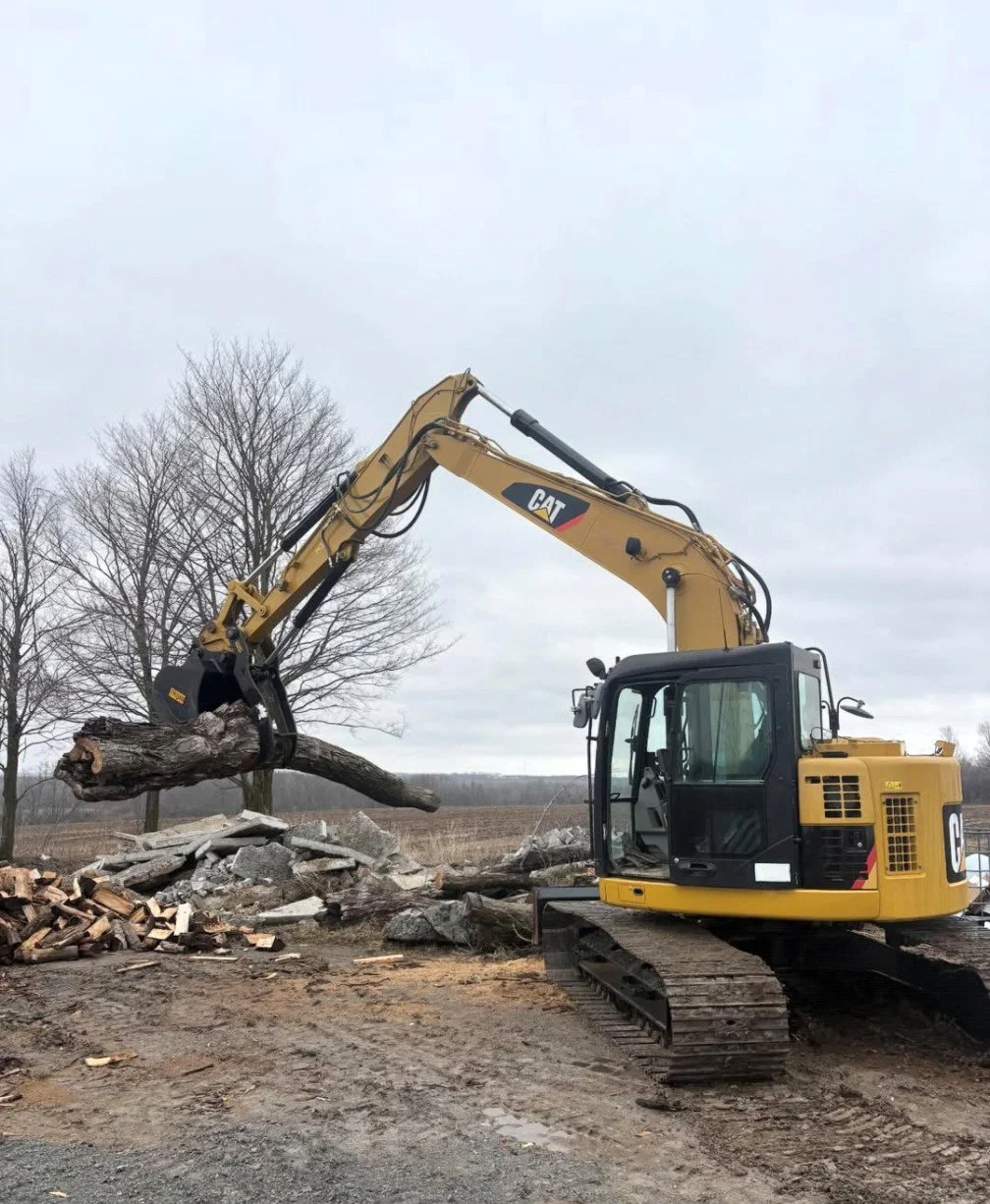 A yellow CAT excavator lifting a large log in a rural outdoor area with barren trees and gray sky.