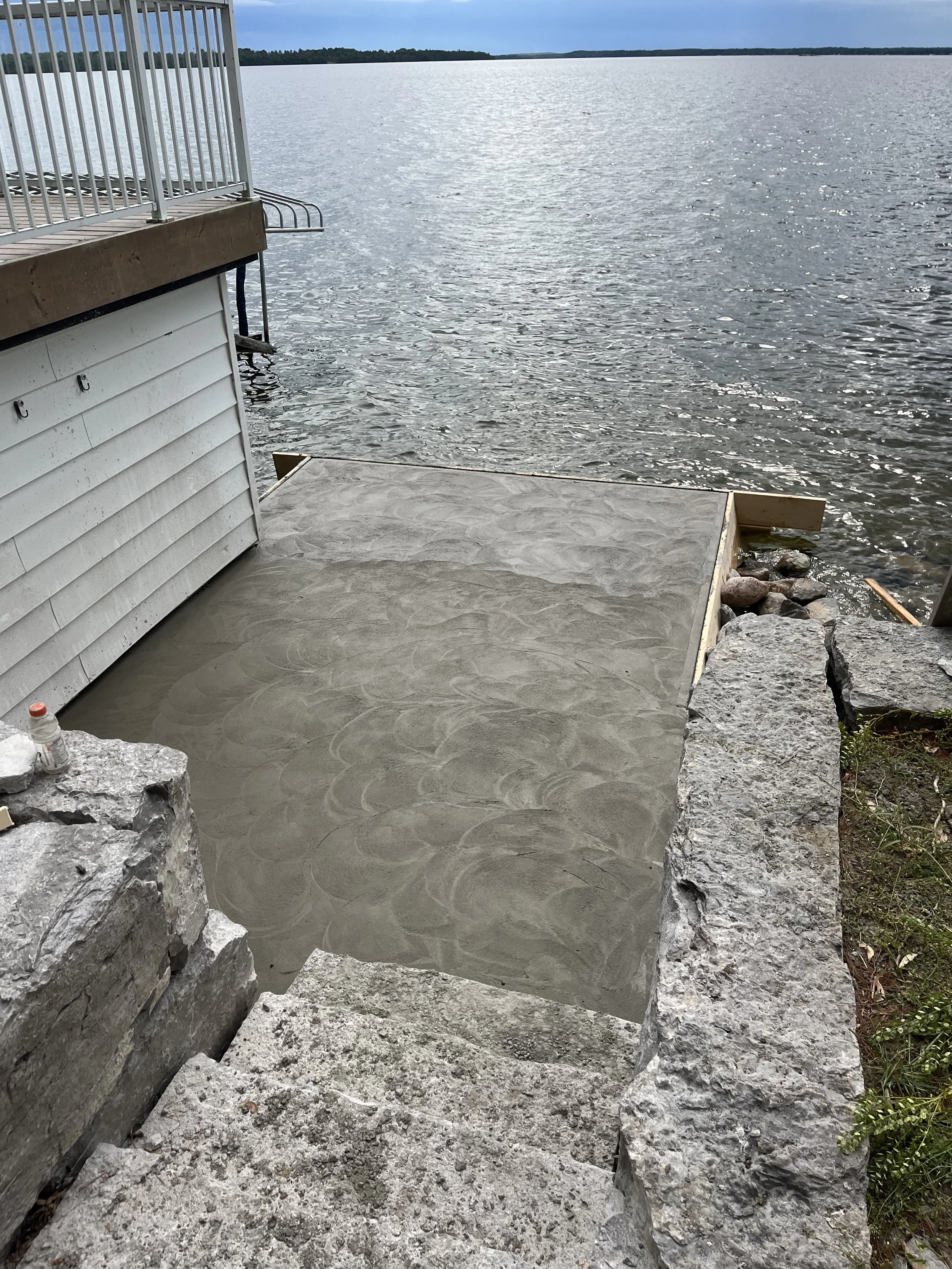 A dock under construction with a concrete platform and rocks surrounding it, overlooking a body of water with a distant treeline and a cloudy sky.