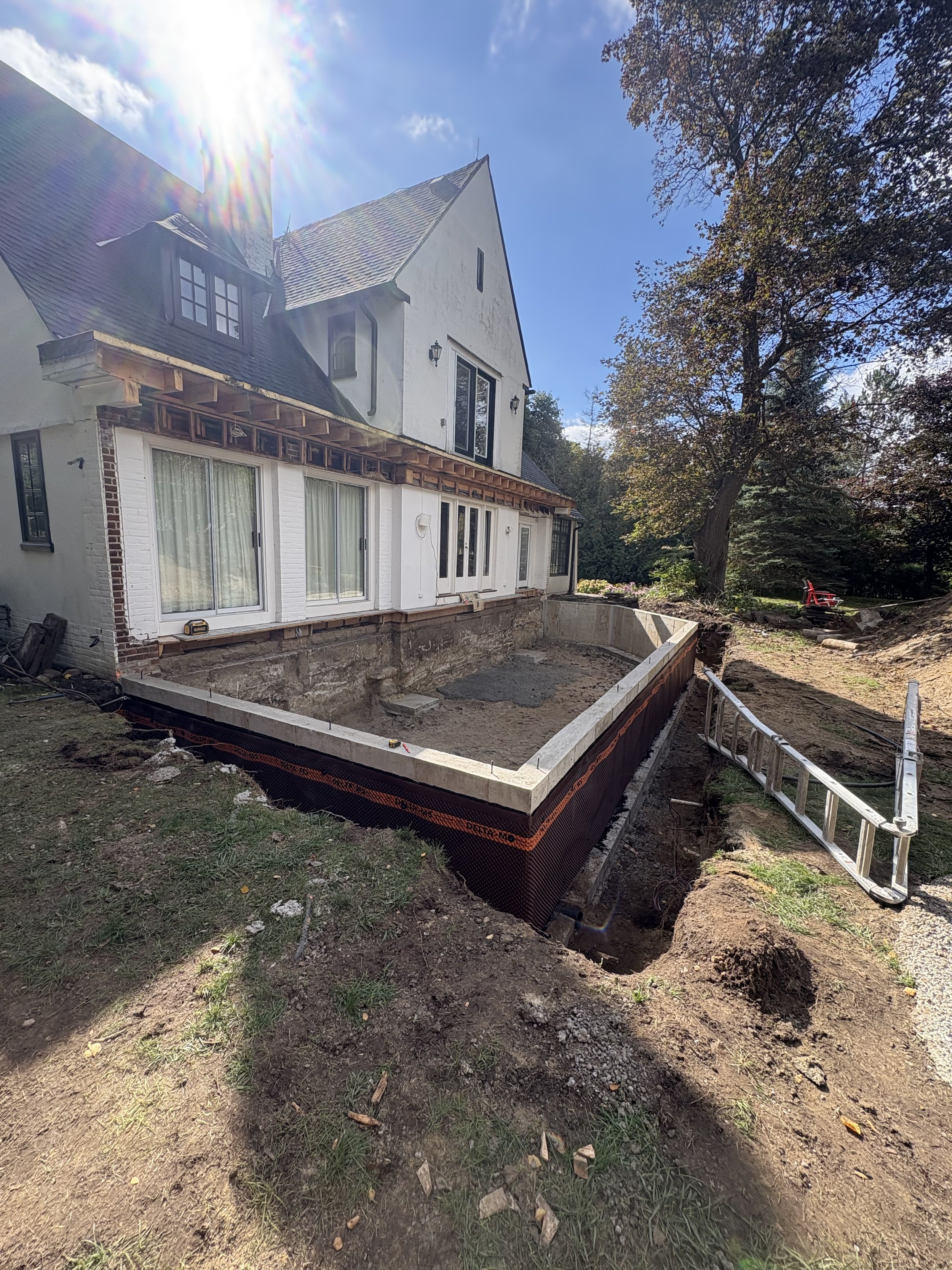A house in Bowmanville under renovation with a partially built extension, exposing the foundation and framing, on a sunny day with clear blue sky and trees nearby.