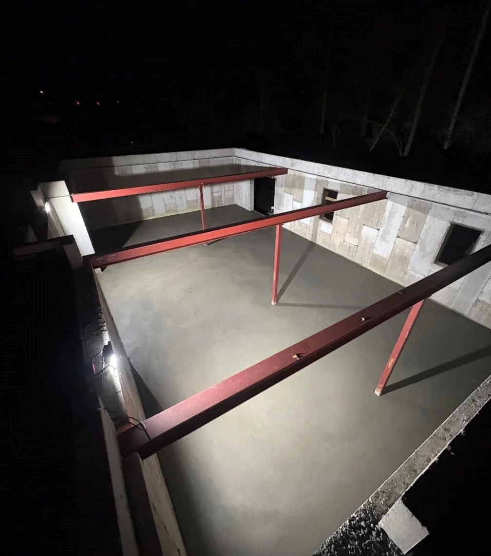 Nighttime view of a construction site showing a concrete foundation with steel beams installed, surrounded by walls made of beige stone blocks.