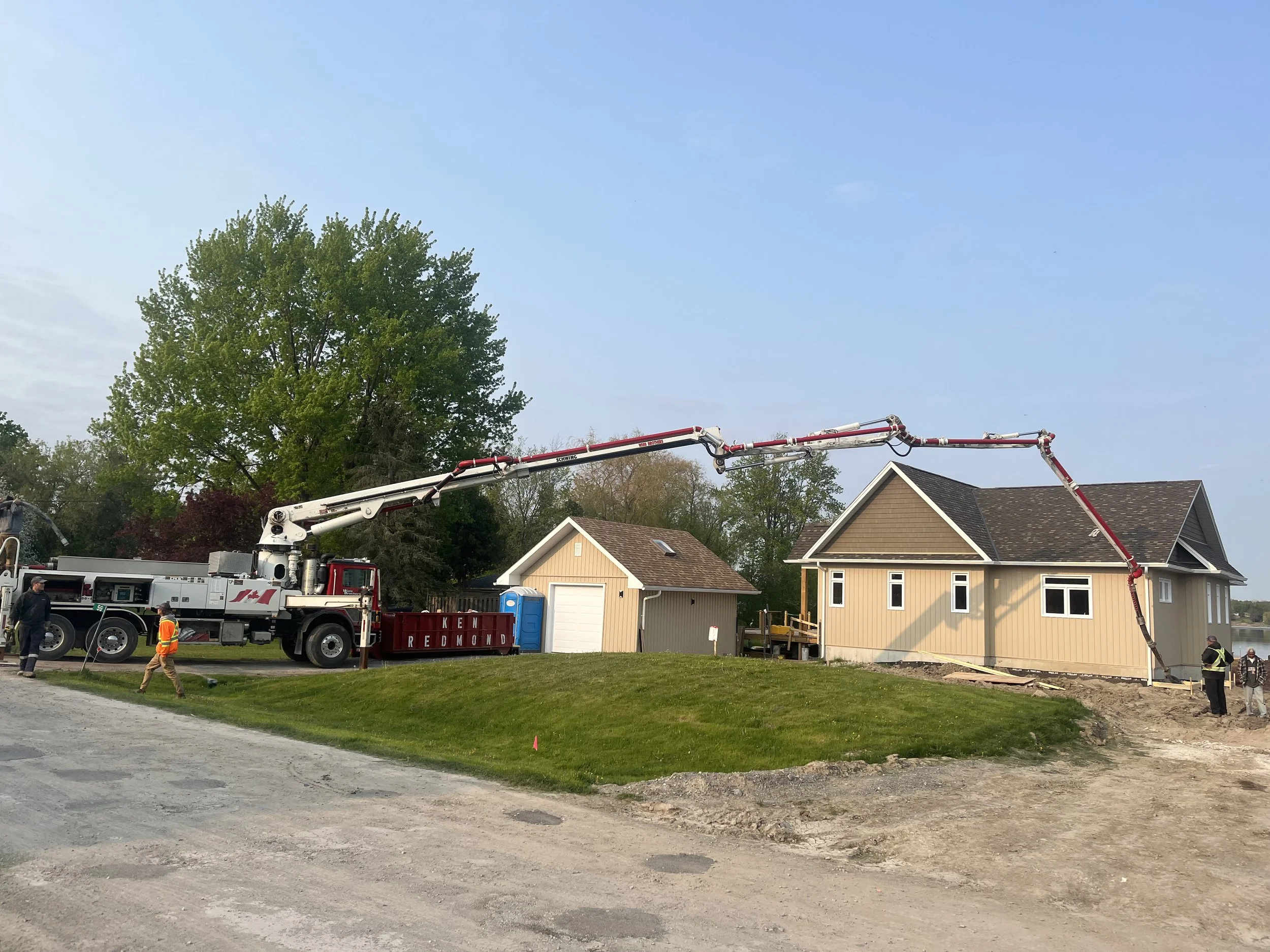 Construction workers and a concrete pump truck pouring foundation at a residential site with houses and trees in the background.
