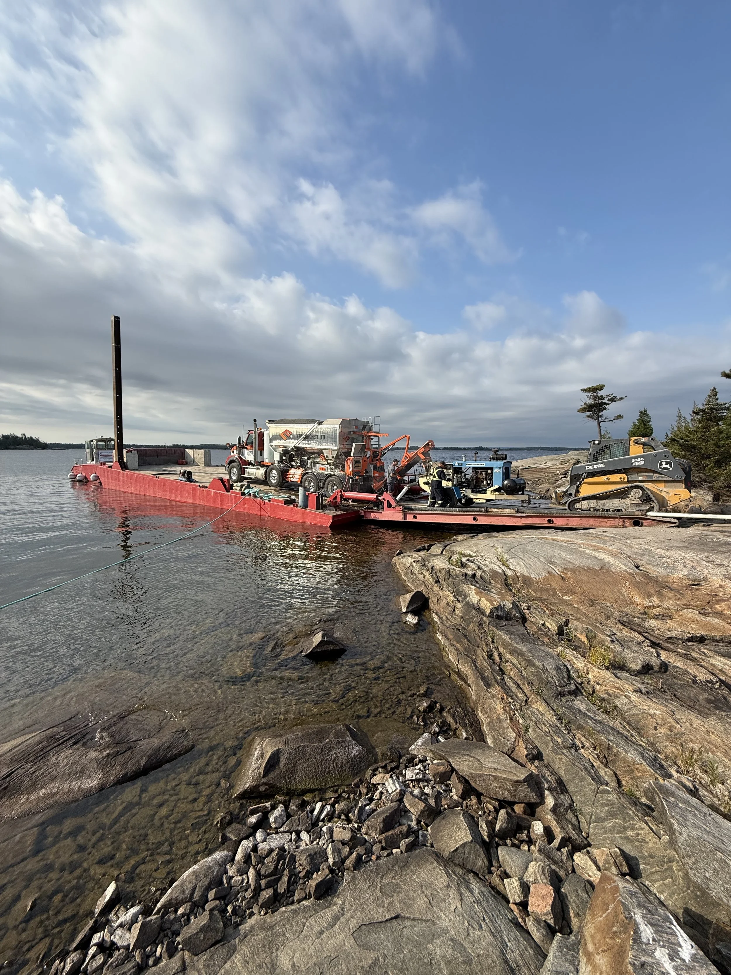 Construction site near water with large red barge, construction vehicles, rocky shoreline, and cloudy sky.