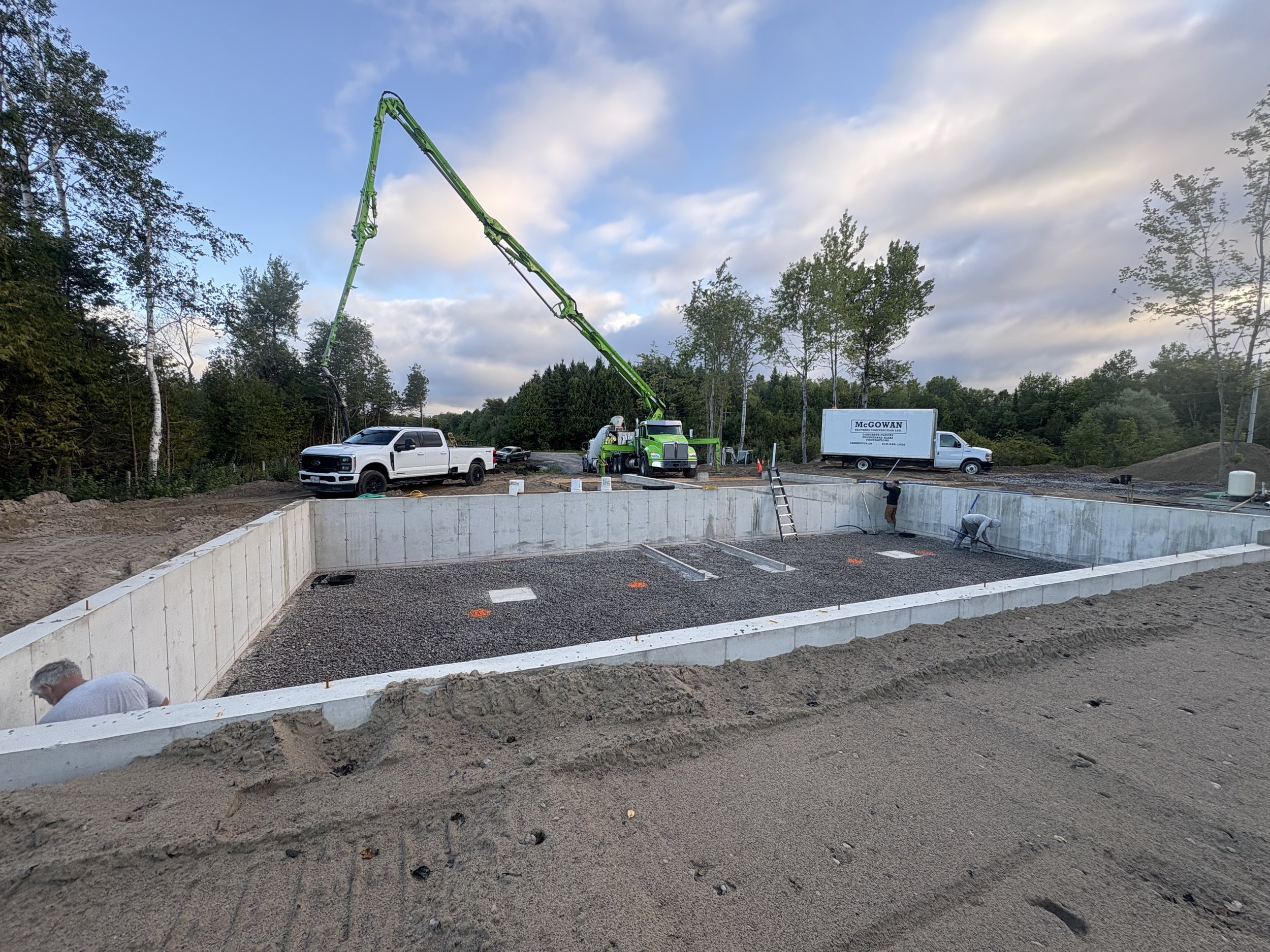 Construction site with a concrete foundation, construction vehicles, workers, and equipment, surrounded by trees and a cloudy sky.