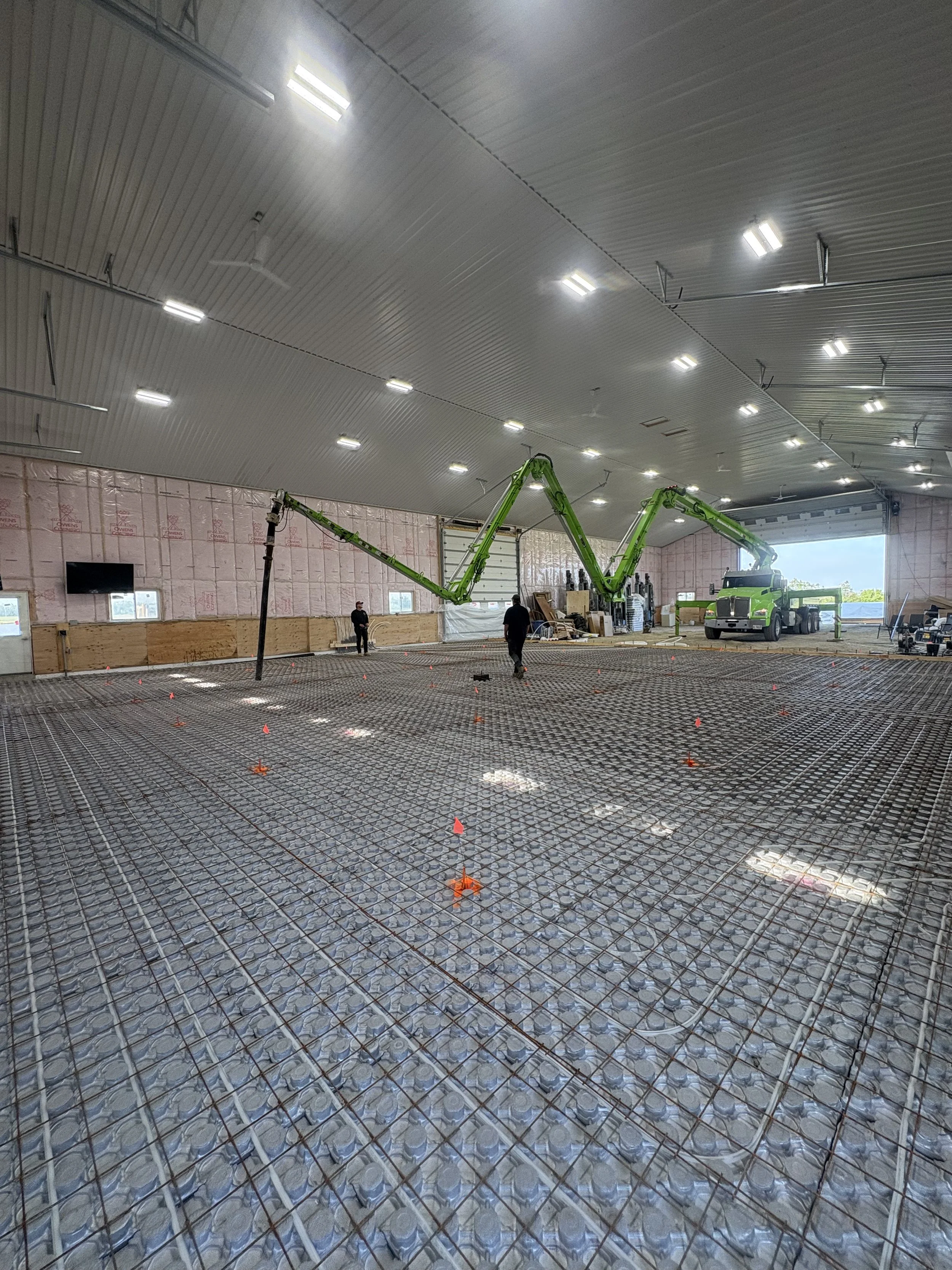 Workers installing steel reinforcement on the floor of a large indoor construction site, with a crane and insulation on the walls.