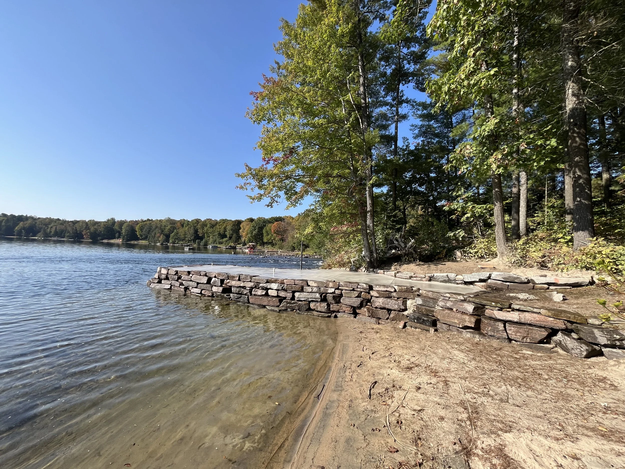 A lakeside scene with a sandy shoreline, clear water, and a small stone wall. In the background, dense trees with green leaves extend to the horizon under a blue sky with no clouds.