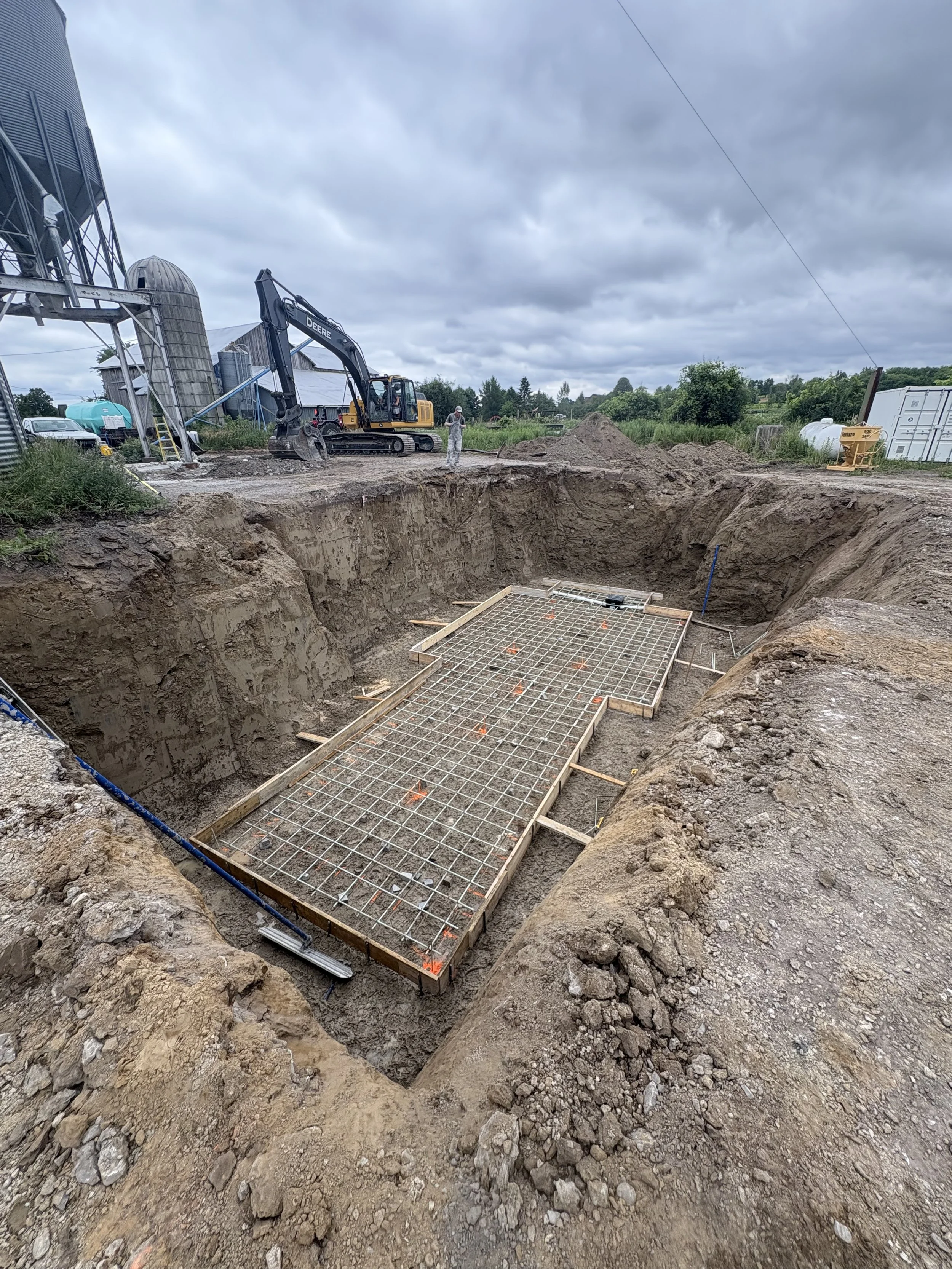 Construction site with an excavator working near a large trench and a metal grid foundation for a building, with cloudy sky and rural surroundings.