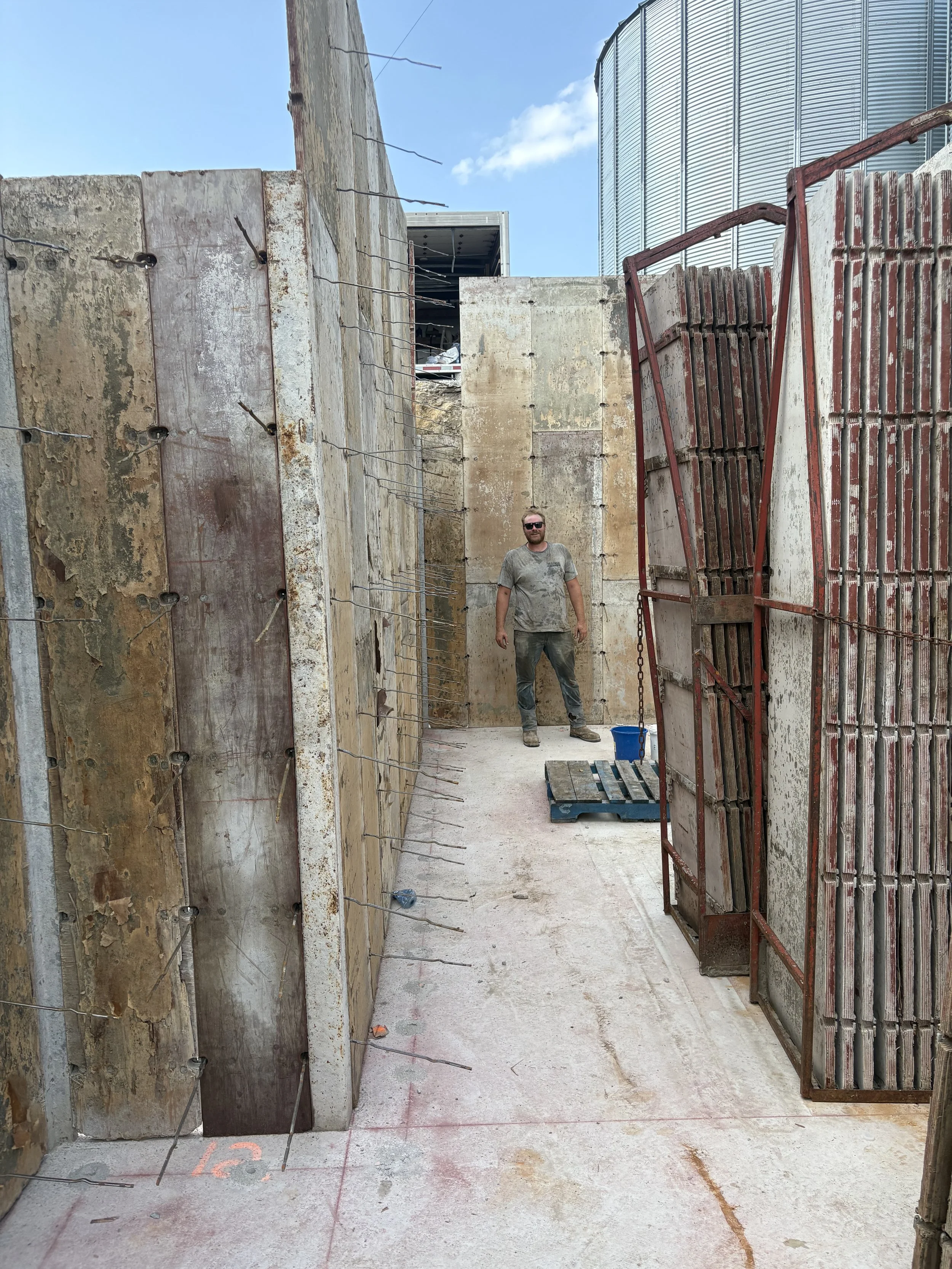 A construction man standing inside a narrow concrete corridor on a construction site. The man is wearing sunglasses, a gray t-shirt, and jeans. There are concrete and steel walls on both sides, some with rust and exposed rebar, and a large metal silo