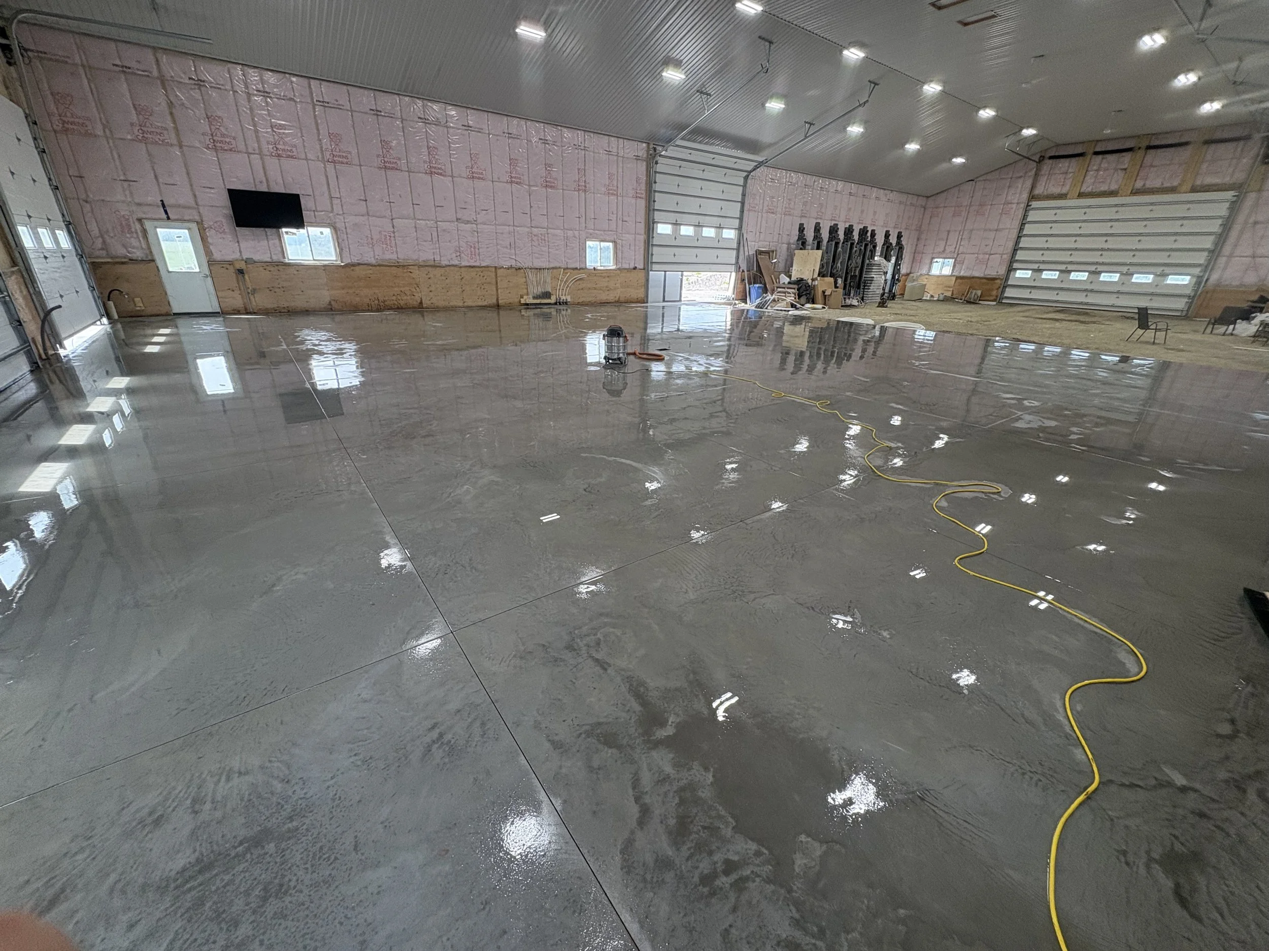A large indoor space under construction with newly poured concrete flooring, construction materials, tools, and equipment scattered around, with insulation on the walls and several small windows.