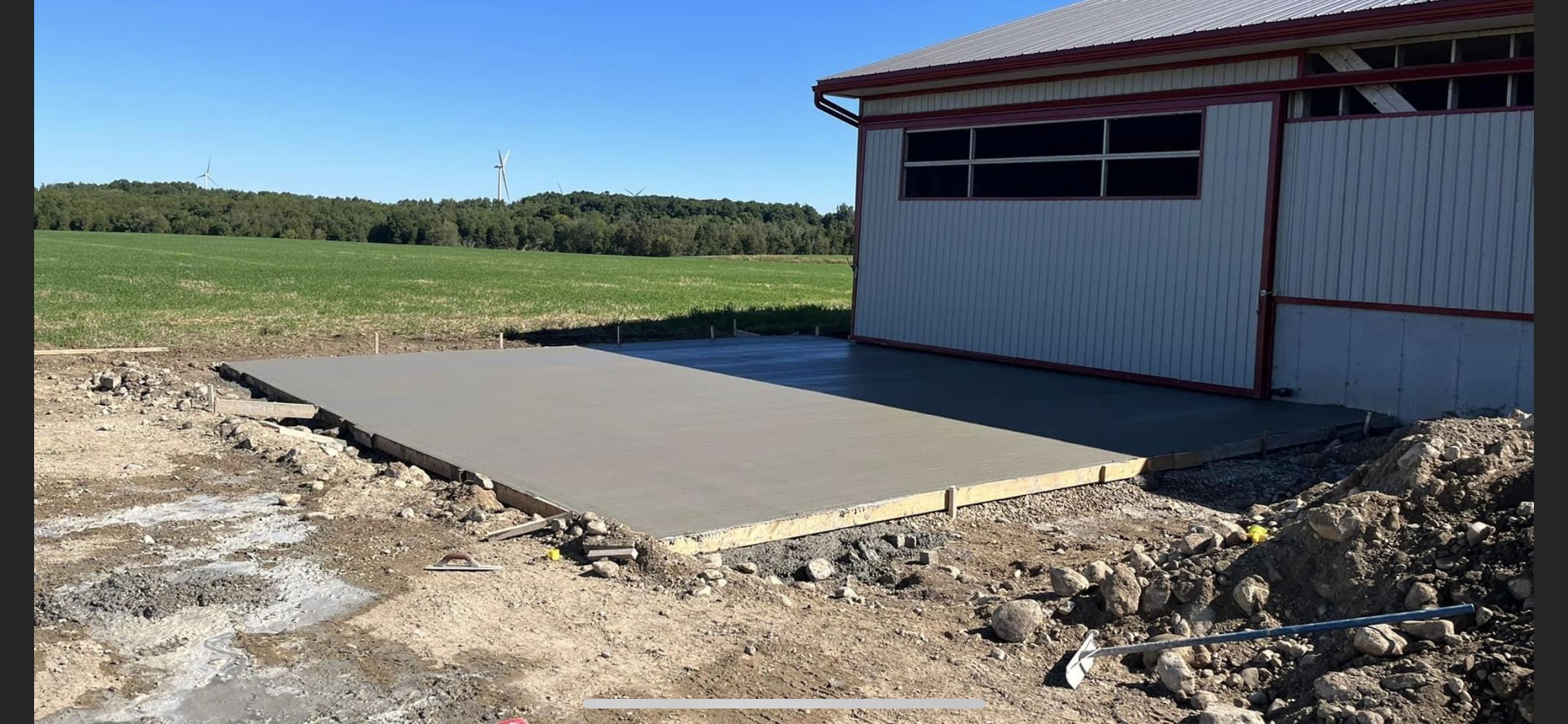 Newly poured concrete slab in front of a partially constructed building on a rural farm, with green fields, trees, and wind turbines in the background.