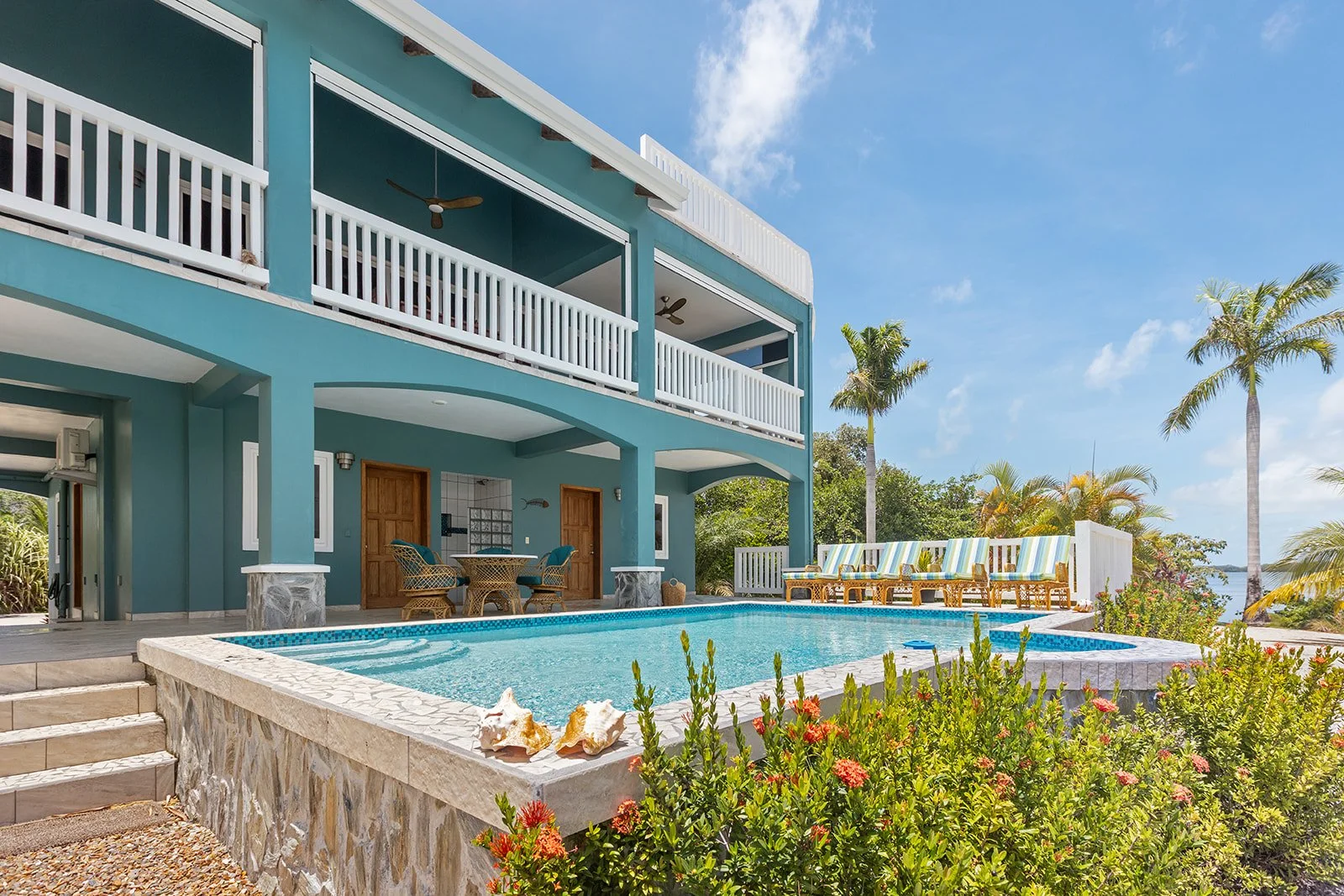 A tropical backyard with a blue two-story house, a swimming pool, palm trees, and outdoor seating on a sunny day.