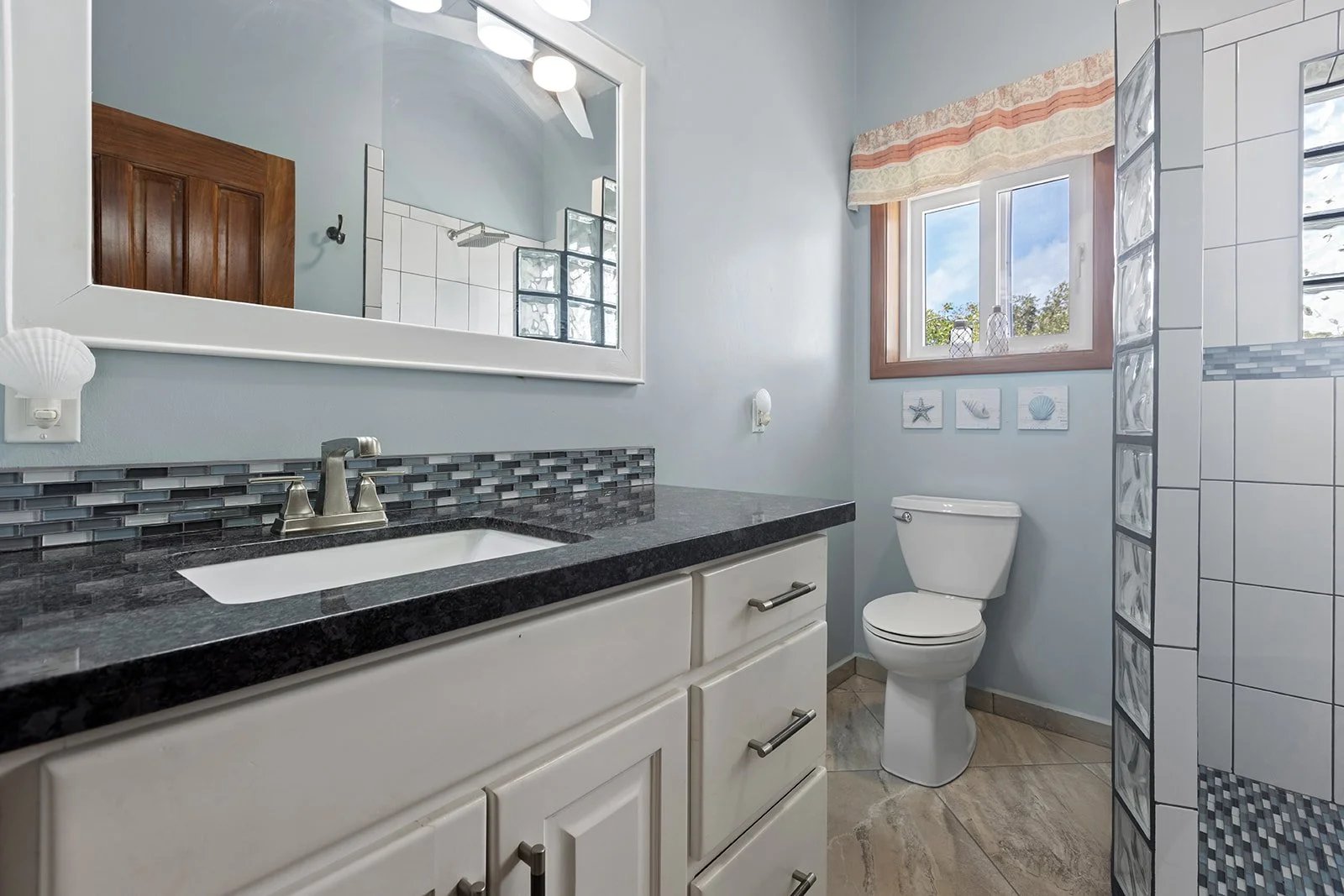 Bathroom with light blue walls, white vanity with black countertop, decorative tile backsplash, large mirror, wall sconce, toilet, window with valance, and shower with glass block wall and white tiles.