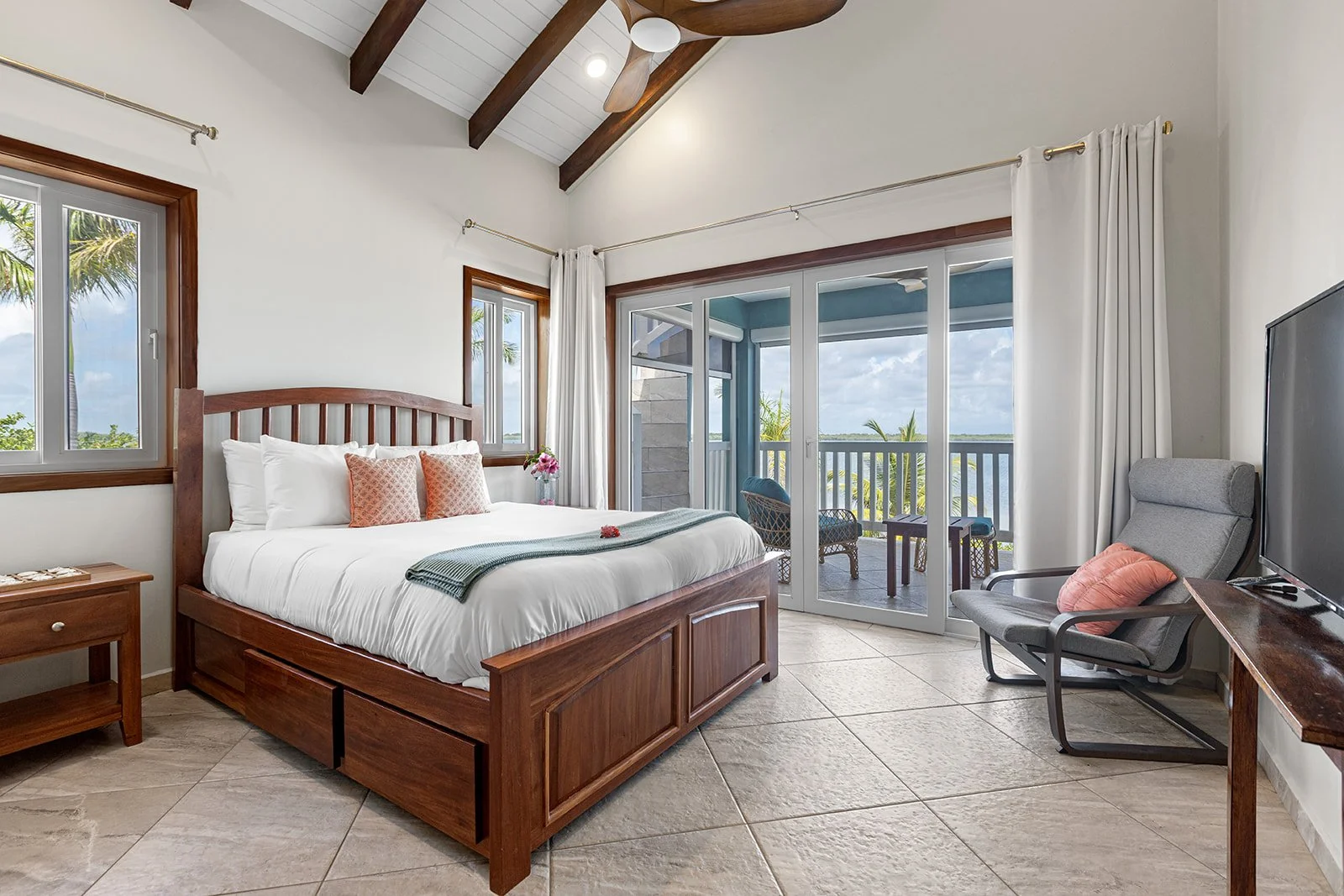 A bedroom with a large bed, two windows, and glass sliding doors leading to a balcony with outdoor furniture, ceiling with wooden beams, and tiled floor.