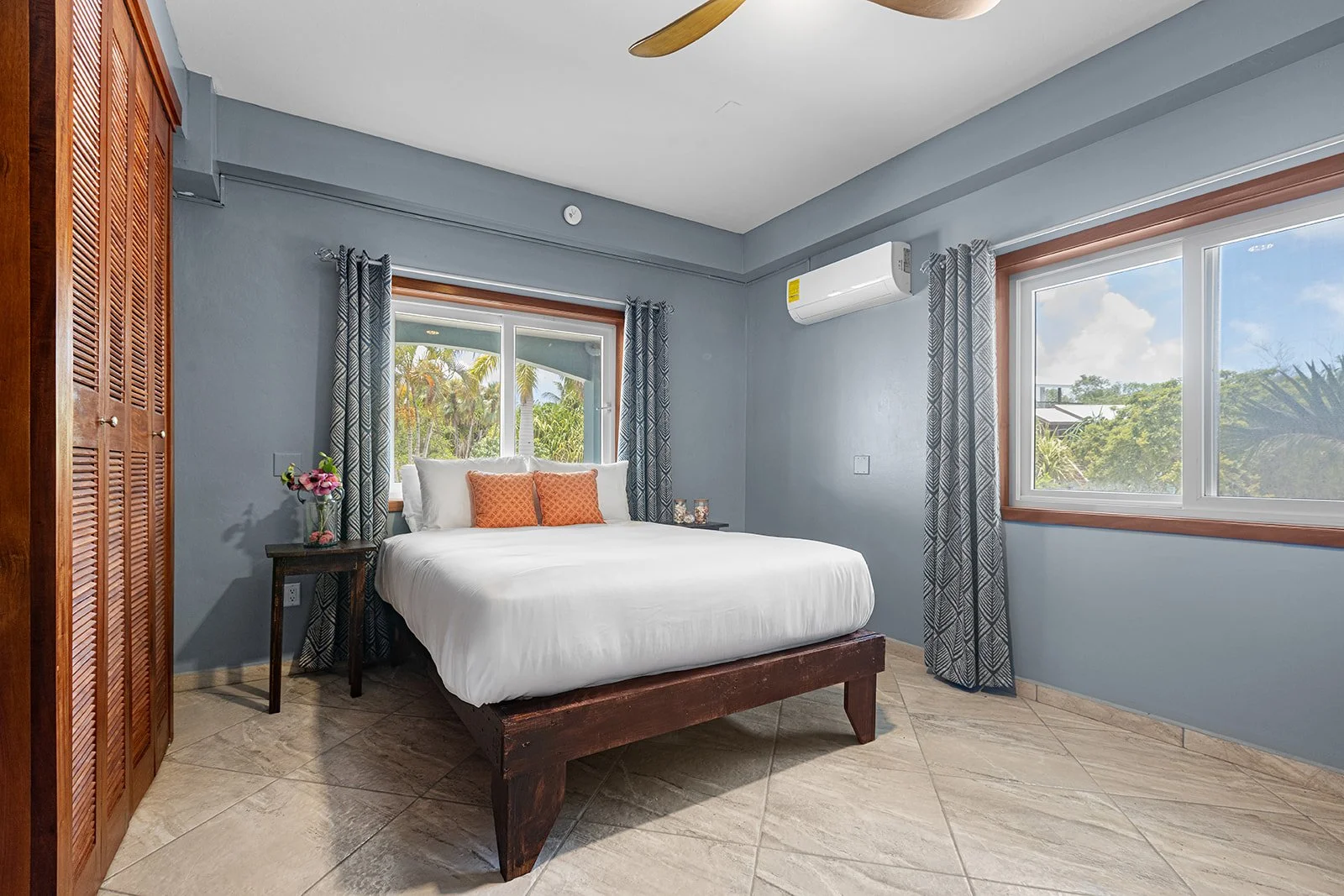 Bedroom with a bed, nightstand with flowers, two windows with curtains, air conditioning unit, and tiled floor.