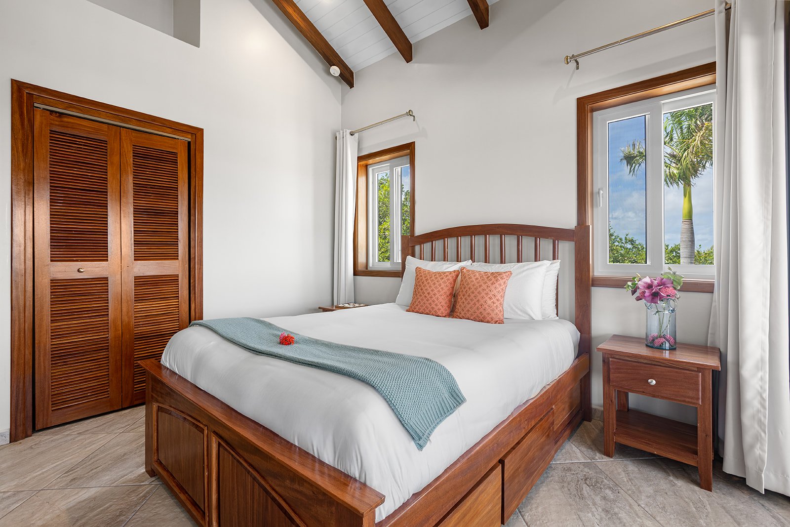 A cozy bedroom with a wooden bed frame, white bedding, two orange pillows, a blue throw blanket, a nightstand with a vase of pink flowers, and two windows showing palm trees outside.