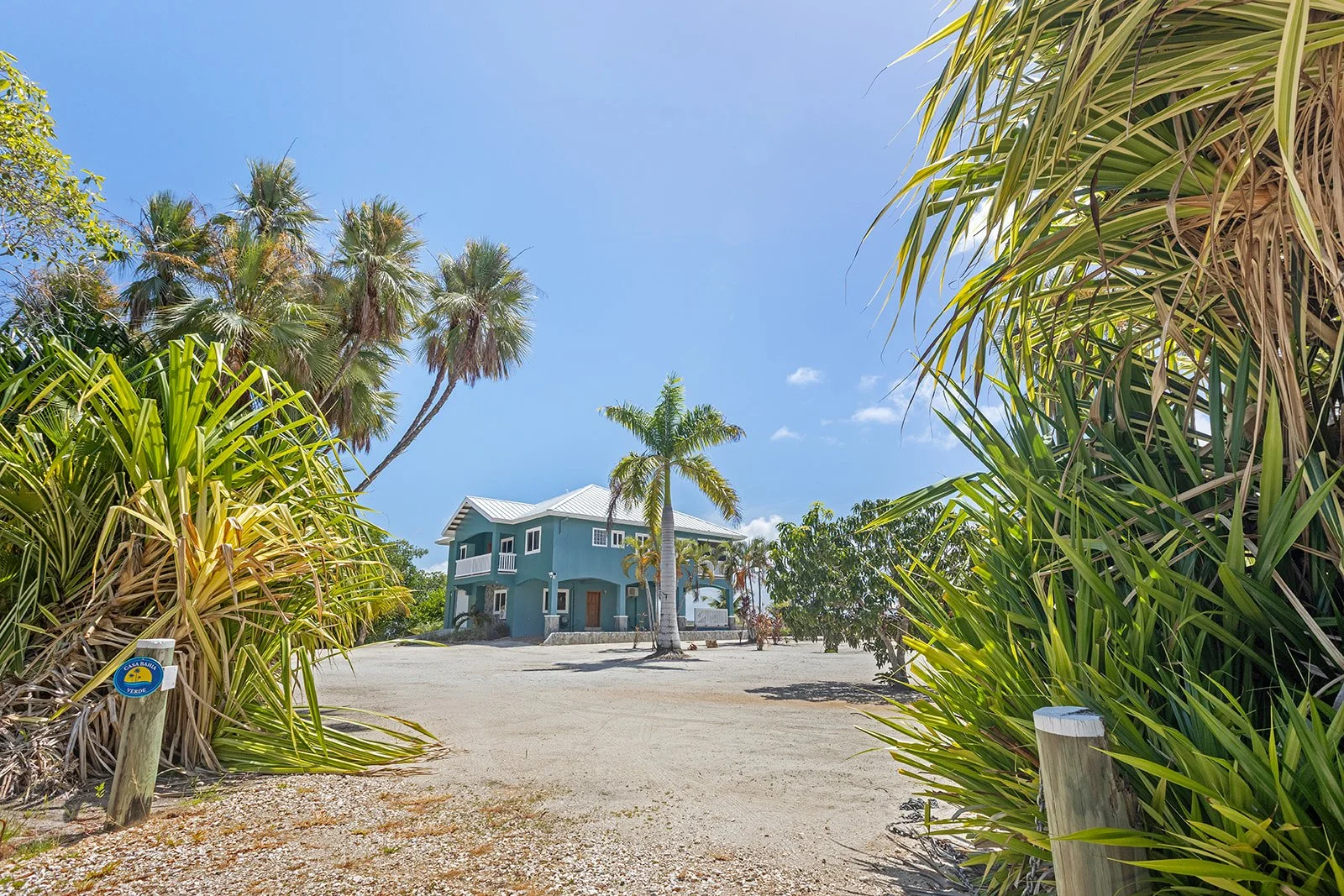 A beach house with blue walls and a white roof surrounded by palm trees and tropical foliage on a sandy beach under a blue sky.