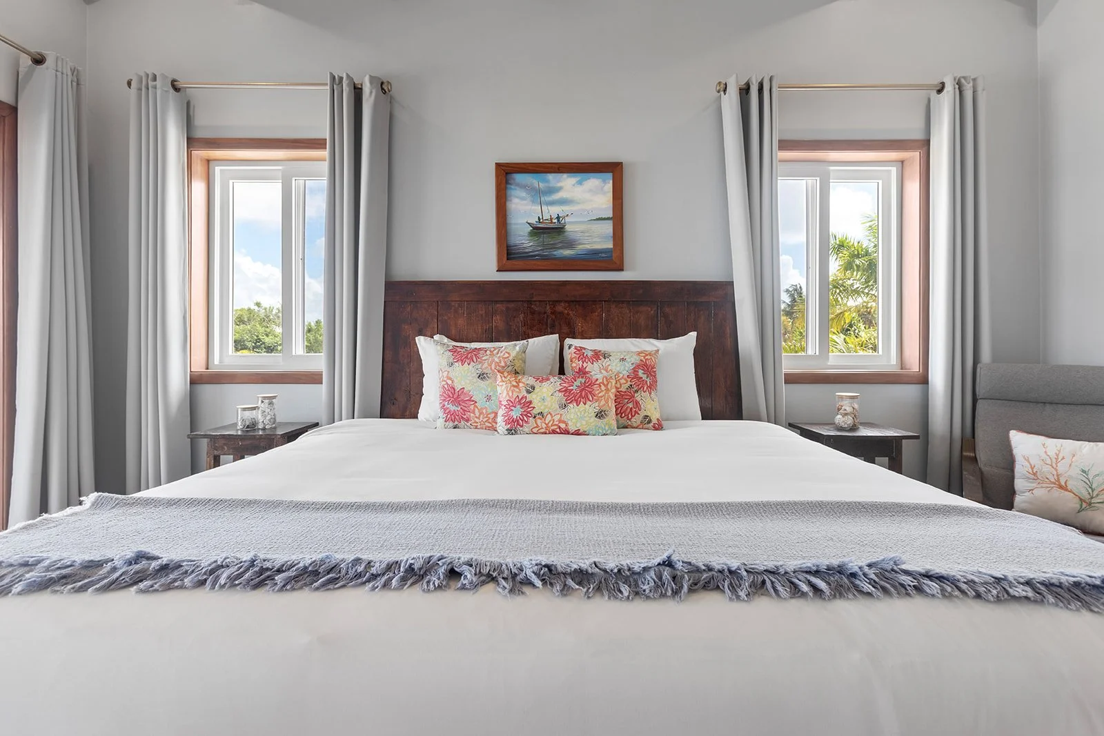A neatly made bed with white linens, decorative pillows with floral patterns, and a throw blanket in a bright, airy bedroom with two windows, curtains, a wooden headboard, and tropical scenery outside.