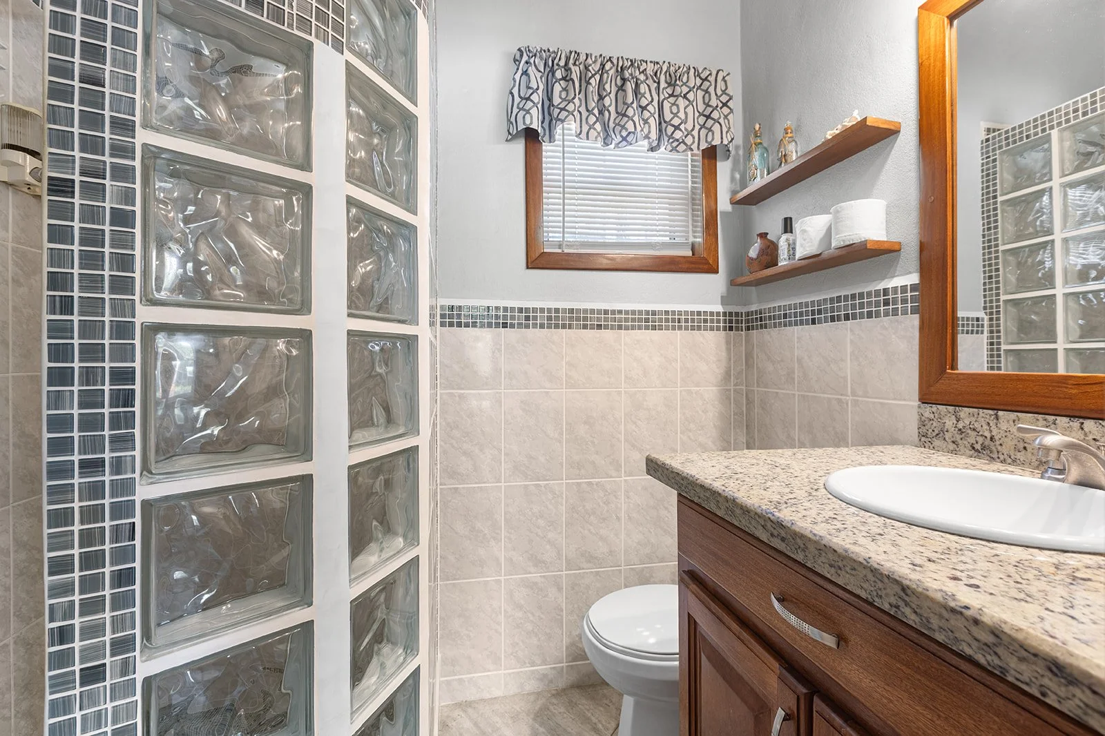 Bathroom with granite countertop vanity, glass block shower wall, small window with valance, wooden shelves with bottles and toilet paper