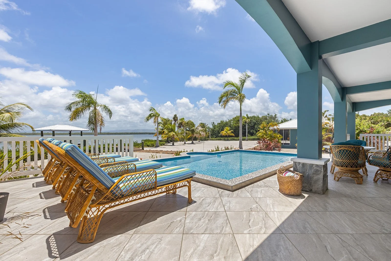 Poolside view with colorful striped lounge chairs, palm trees, and a view of water under a blue sky with scattered clouds.