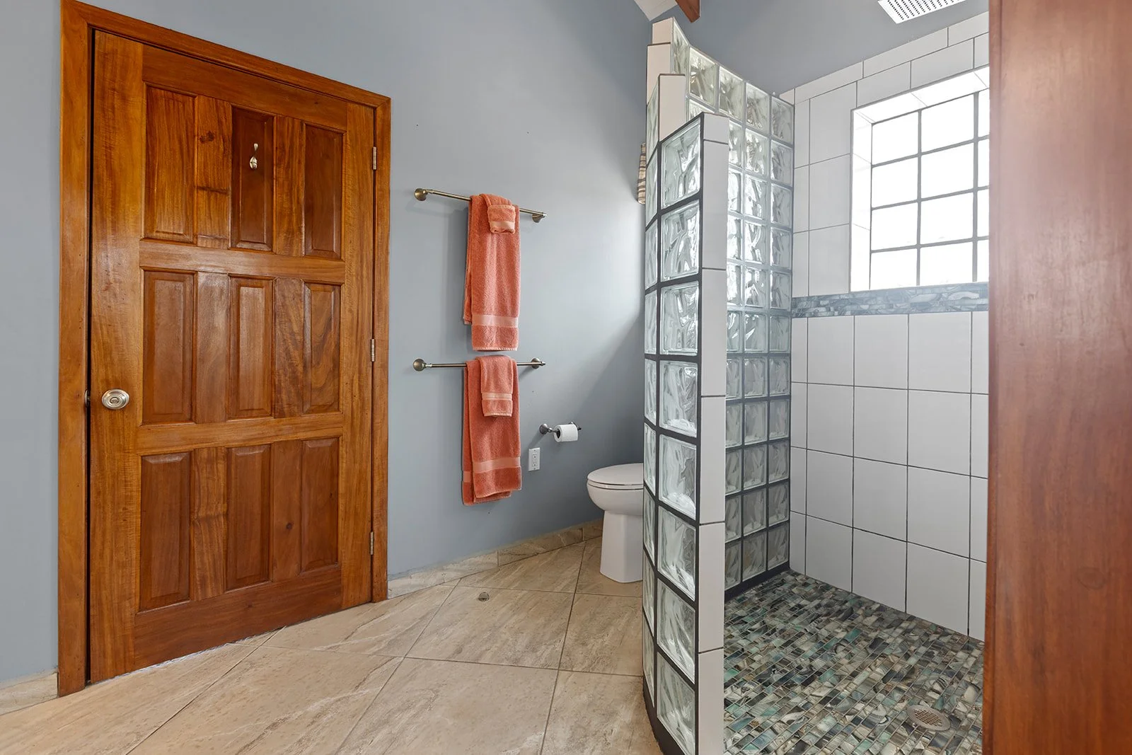 Bathroom with wooden door, two orange towels on a towel rack, toilet, toilet paper holder, frosted glass block shower with white tiles, and a large window.