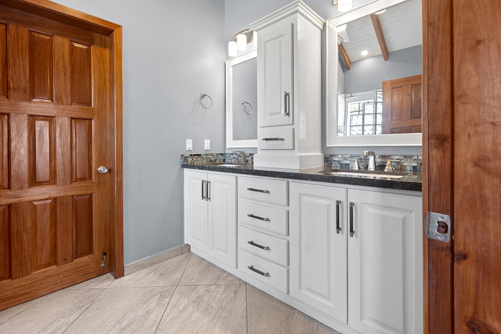 Bathroom vanity with white cabinets, black countertop, and large mirror, wood door on the left, gray wall, tiled floor.