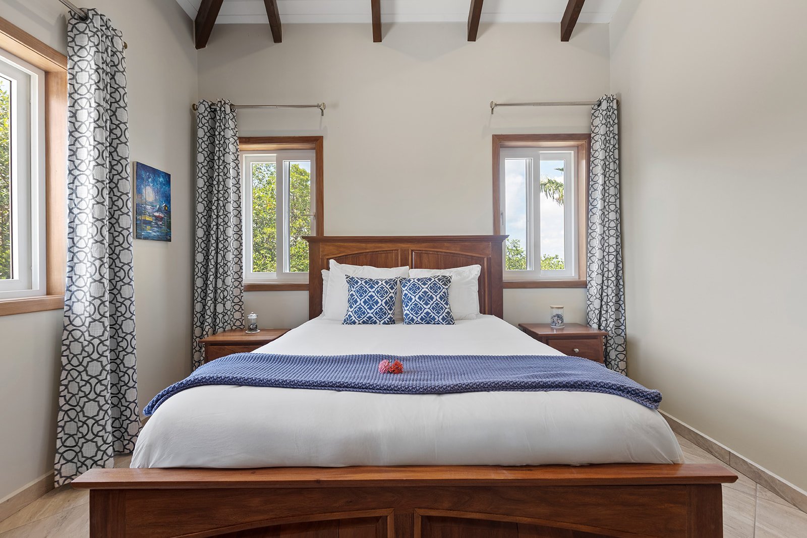 A bedroom with a wooden bed frame, white bedding, blue accent pillows, a blue throw blanket, and two nightstands. There are three windows with wooden frames, patterned curtains, and trees visible outside. A small painting hangs on the left wall.
